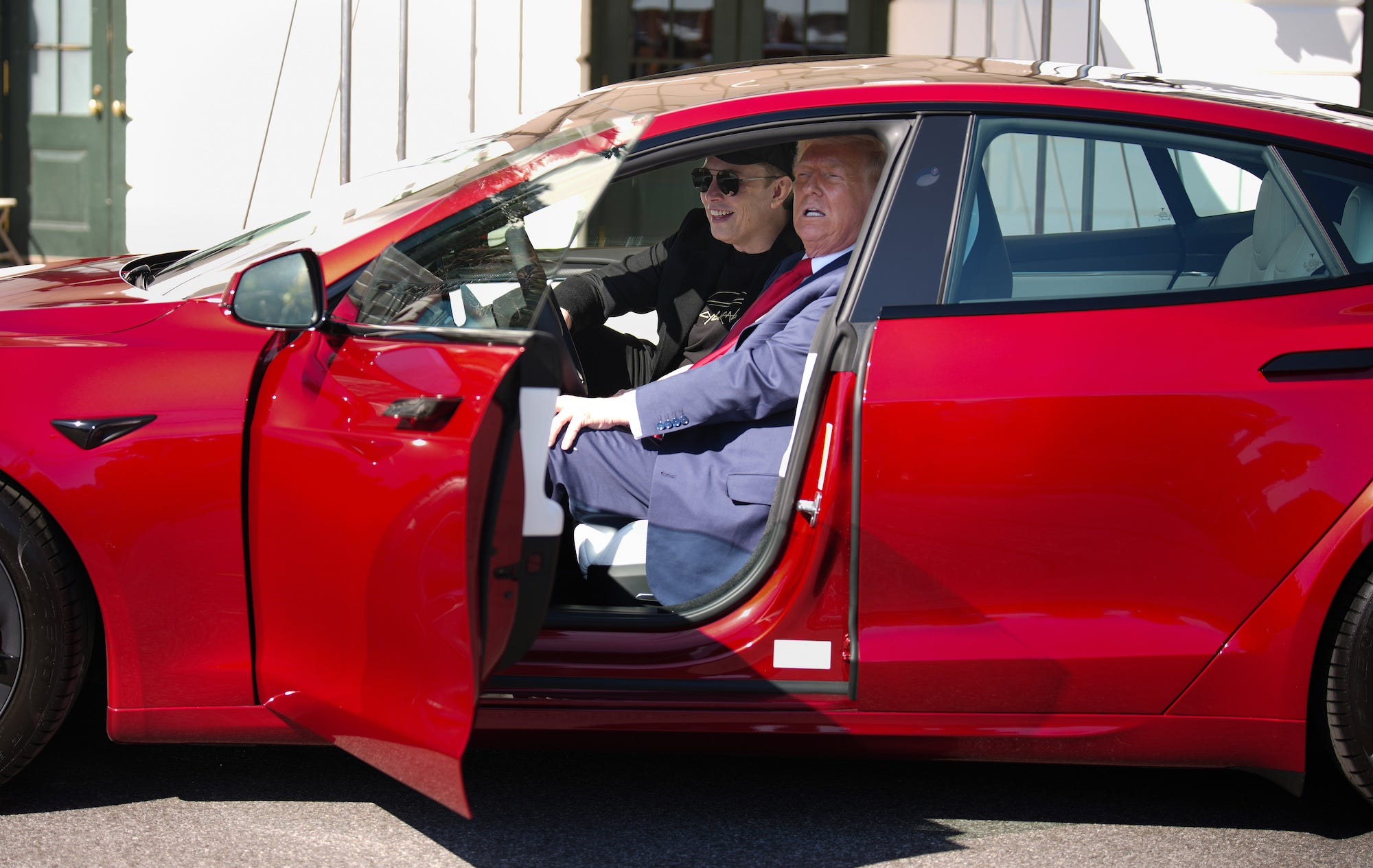 Elon Musk and Donald Trump inside a red Tesla Model S with the door open.