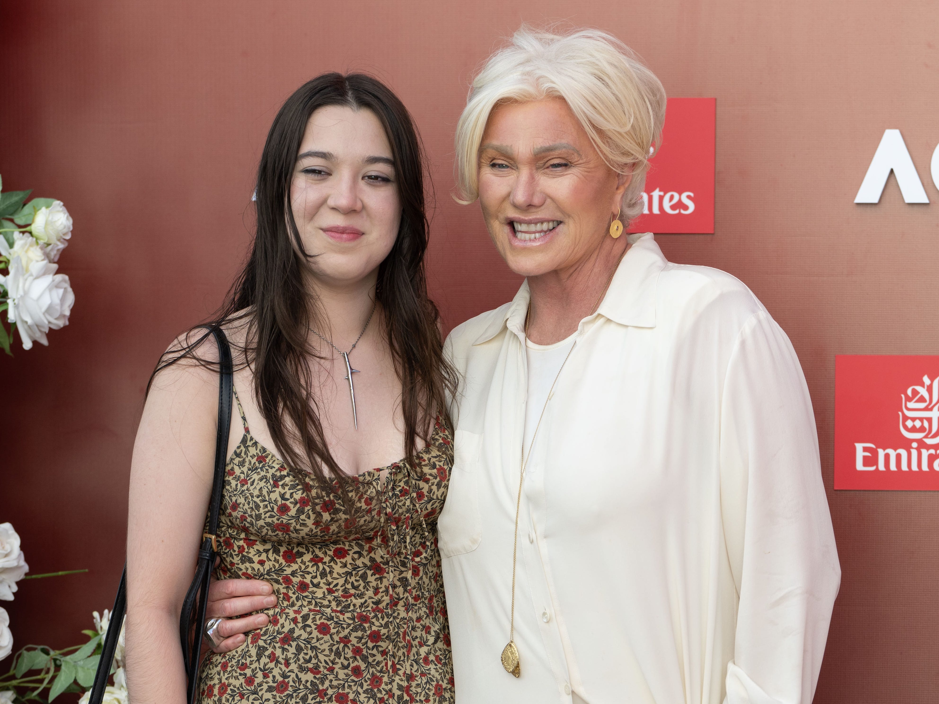 Two women stood on a red carpet. On the left, the younger womn has long brown hair and is wearing a red and brown floral dress. She is also wearing a silver necklace. On the right is an older woman with blond hair who is wearing a flowing white shirt with a gold necklace.