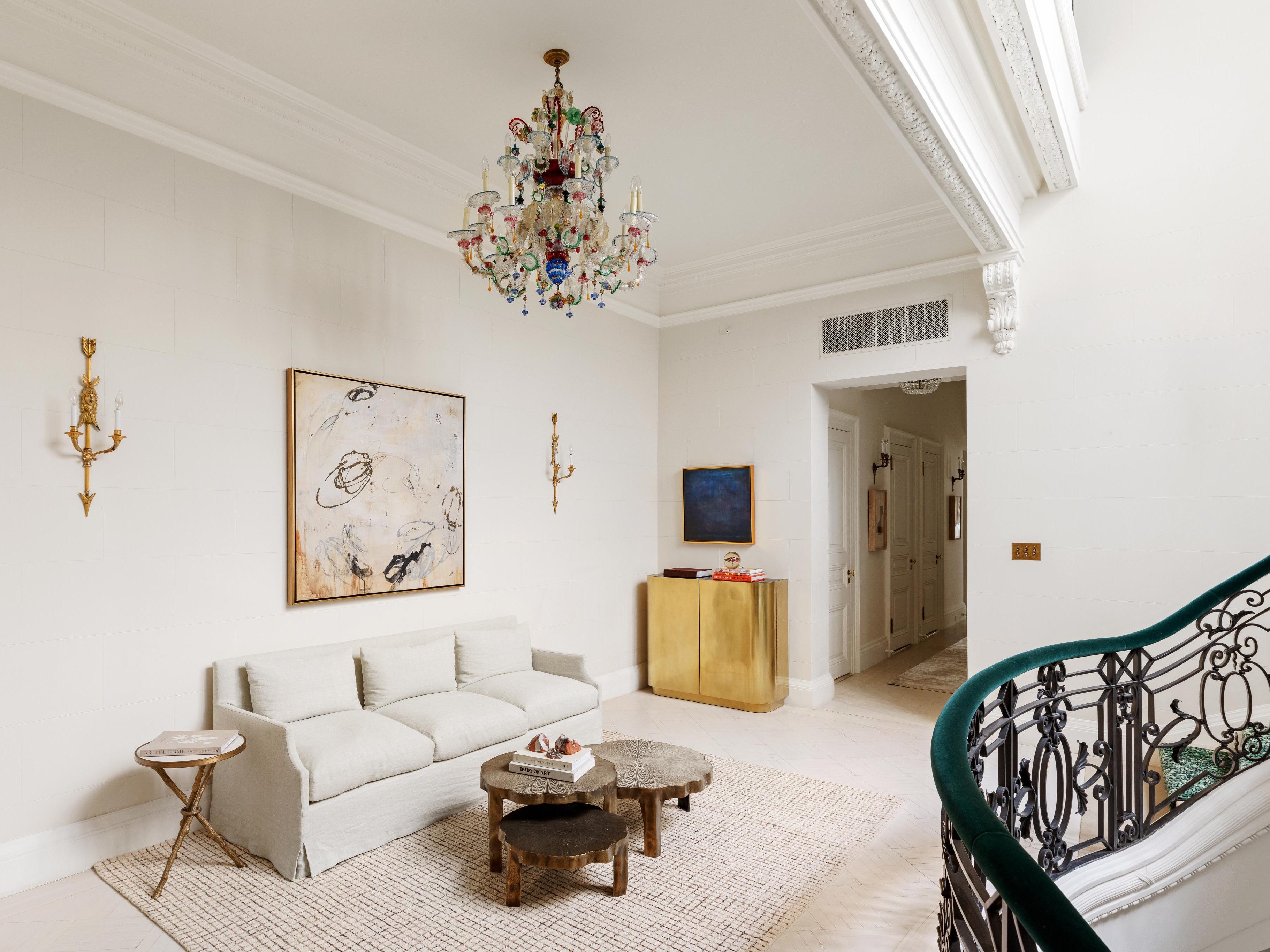 a room opposite a grand staircase. its painted white with a beige couch, chandelier, and modern art on the walls