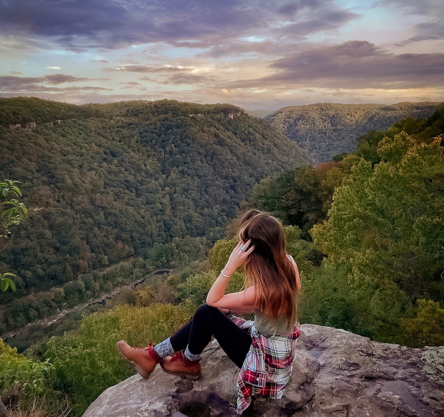 Emily, wearing a green top, a flannel around her waist, black leggings, high socks, and hiking boots, sits on a mountain looking out at the trees.