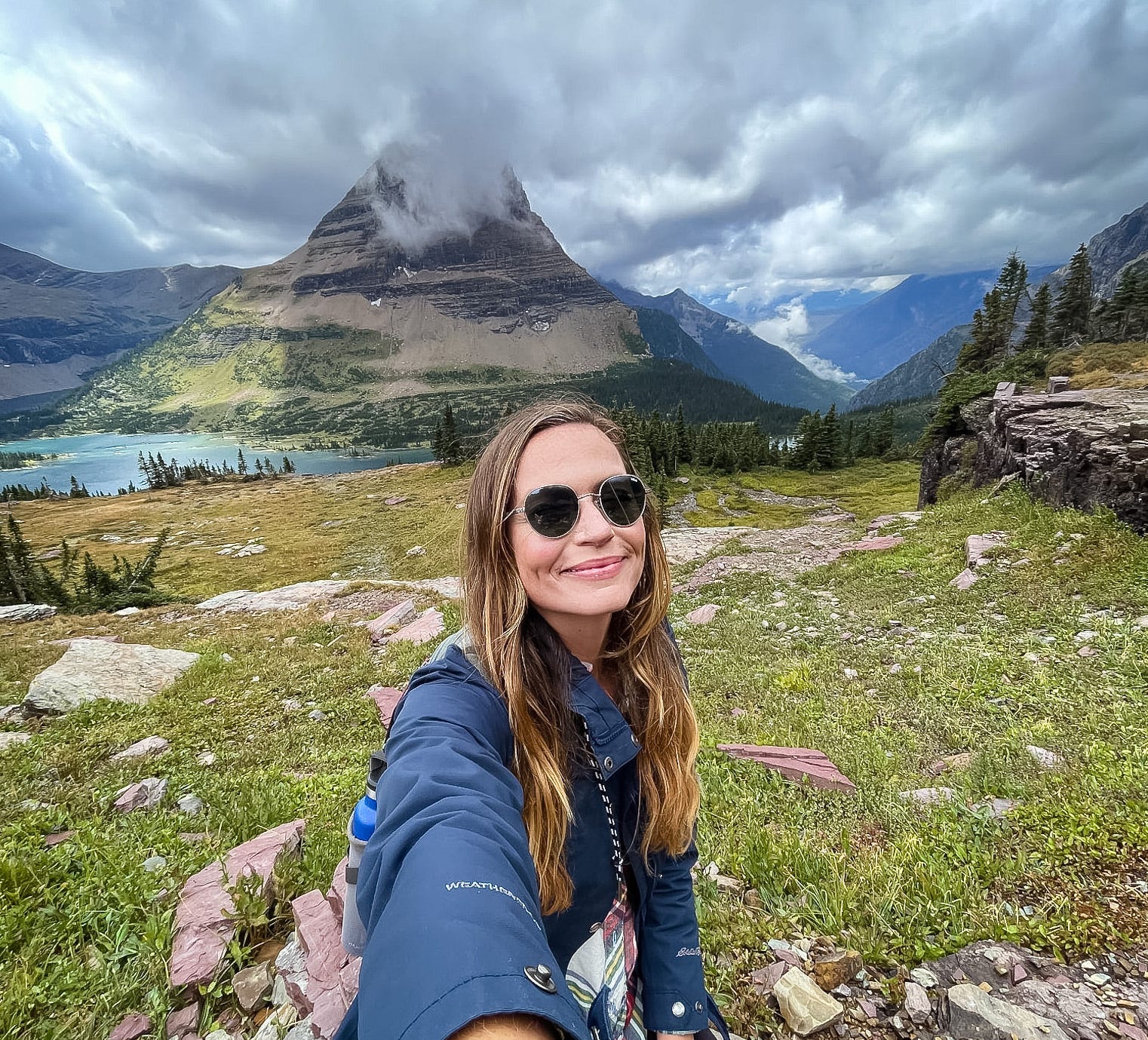 Emily, wearing a blue jacket and sunglasses, poses in a grassy field in front of mountains that stretch into the clouds.