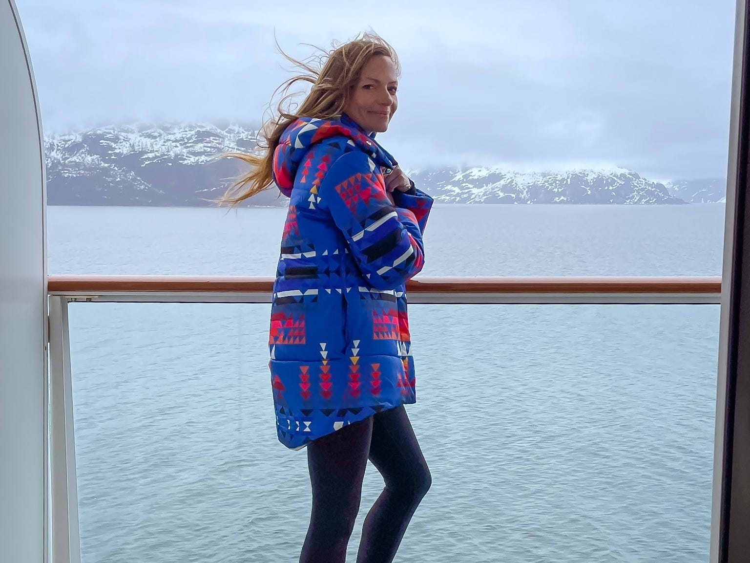 Emily stands on a cruise balcony overlooking glaciers as she wears a heavy coat, black leggings, and grey socks.