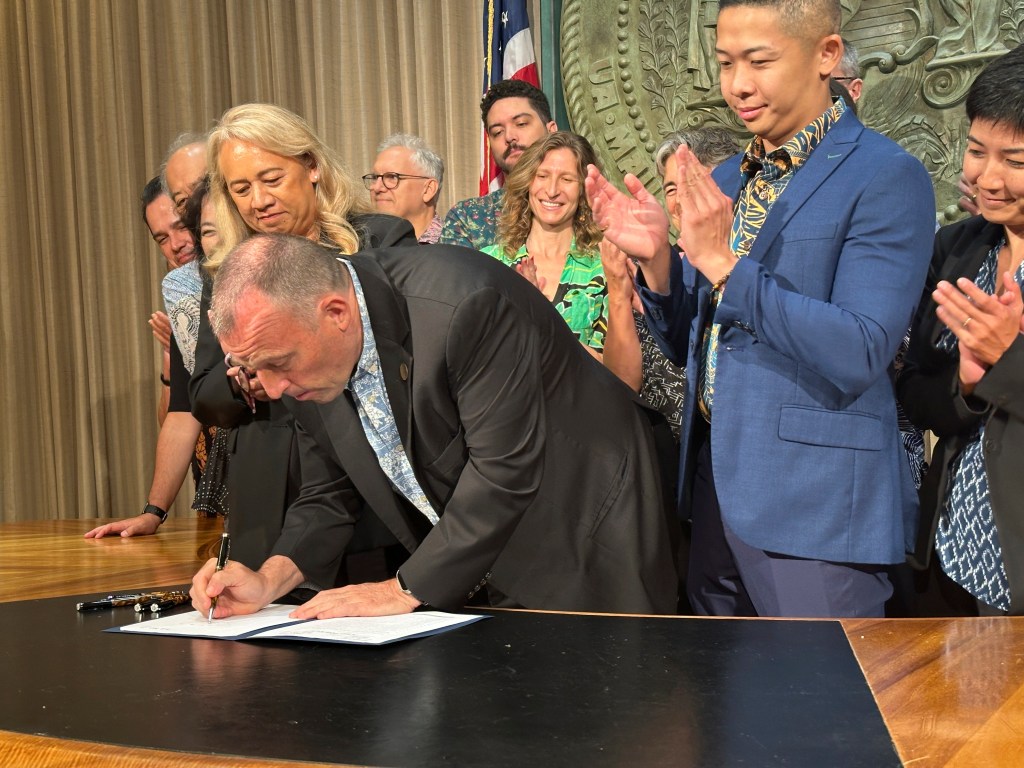 Hawaii Governor Josh Green signing legislation to raise the state tax on short-term accommodations, surrounded by lawmakers and supporters, in an effort to combat climate change