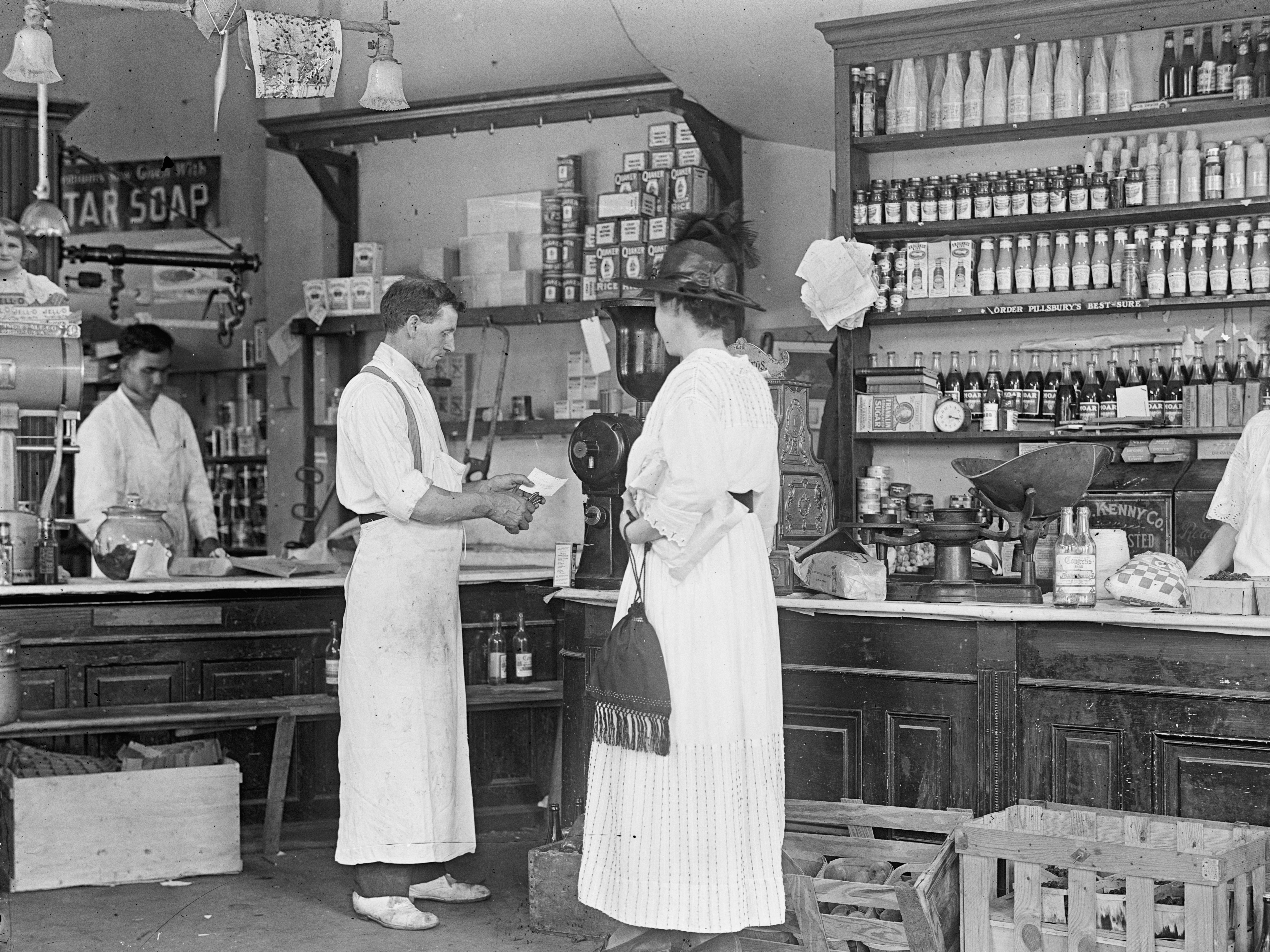 A woman in a long dress is helped by a clerk in a general store