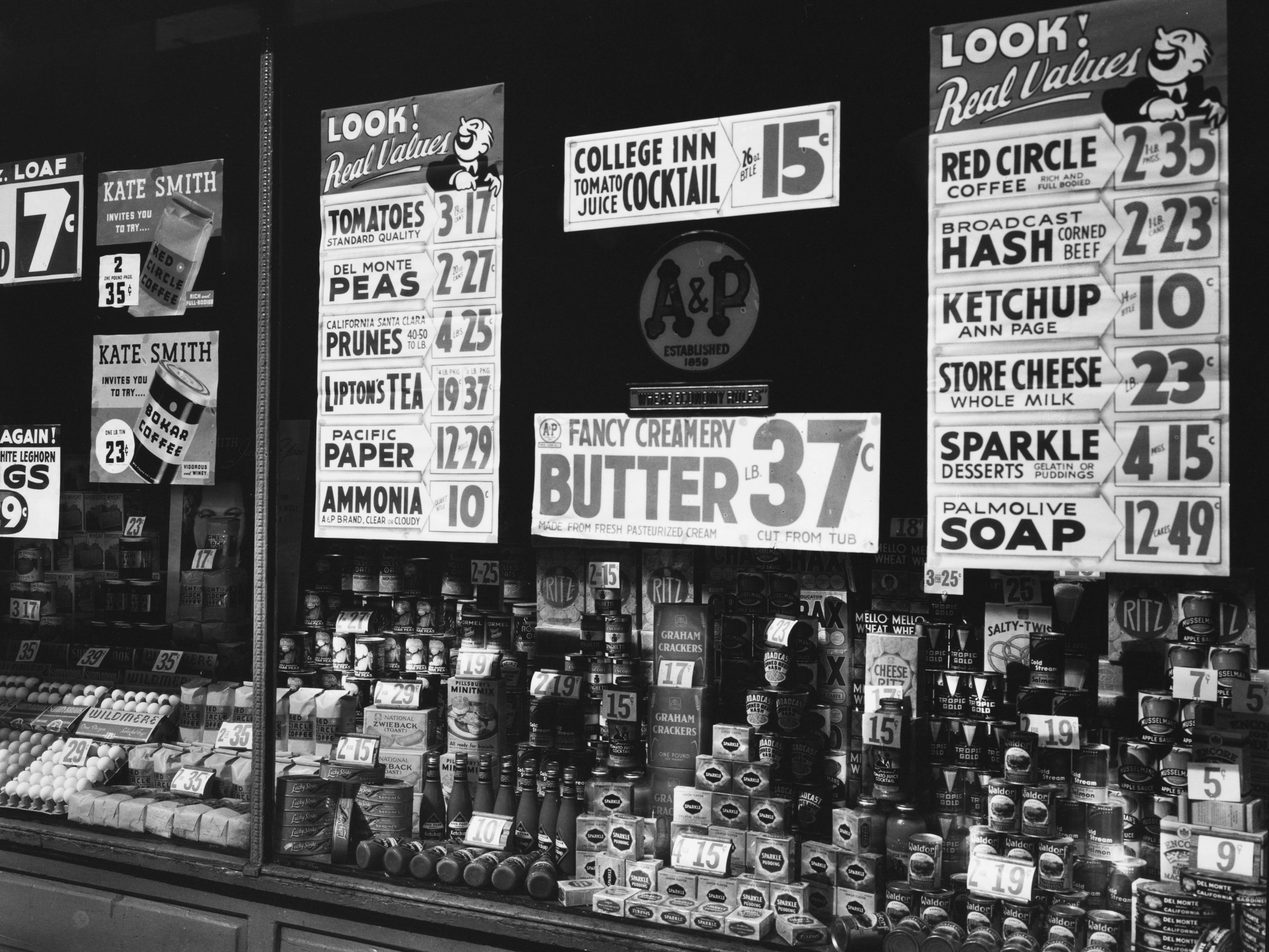 A window with goods stacked up and ads for prunes, amonia, hash, ketchup, and other goods