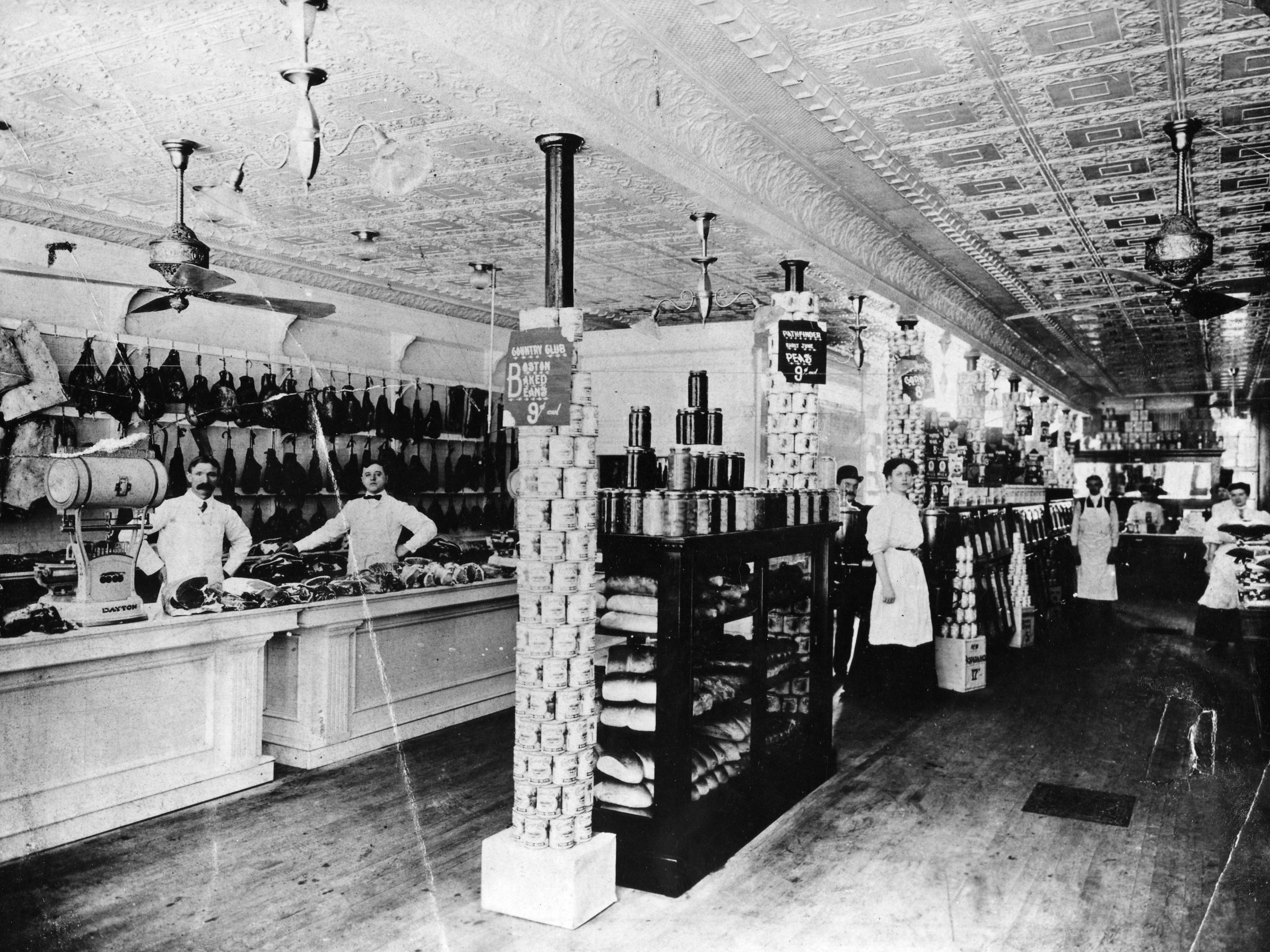 Several people stand in a general store with goods stacked up and on shelves