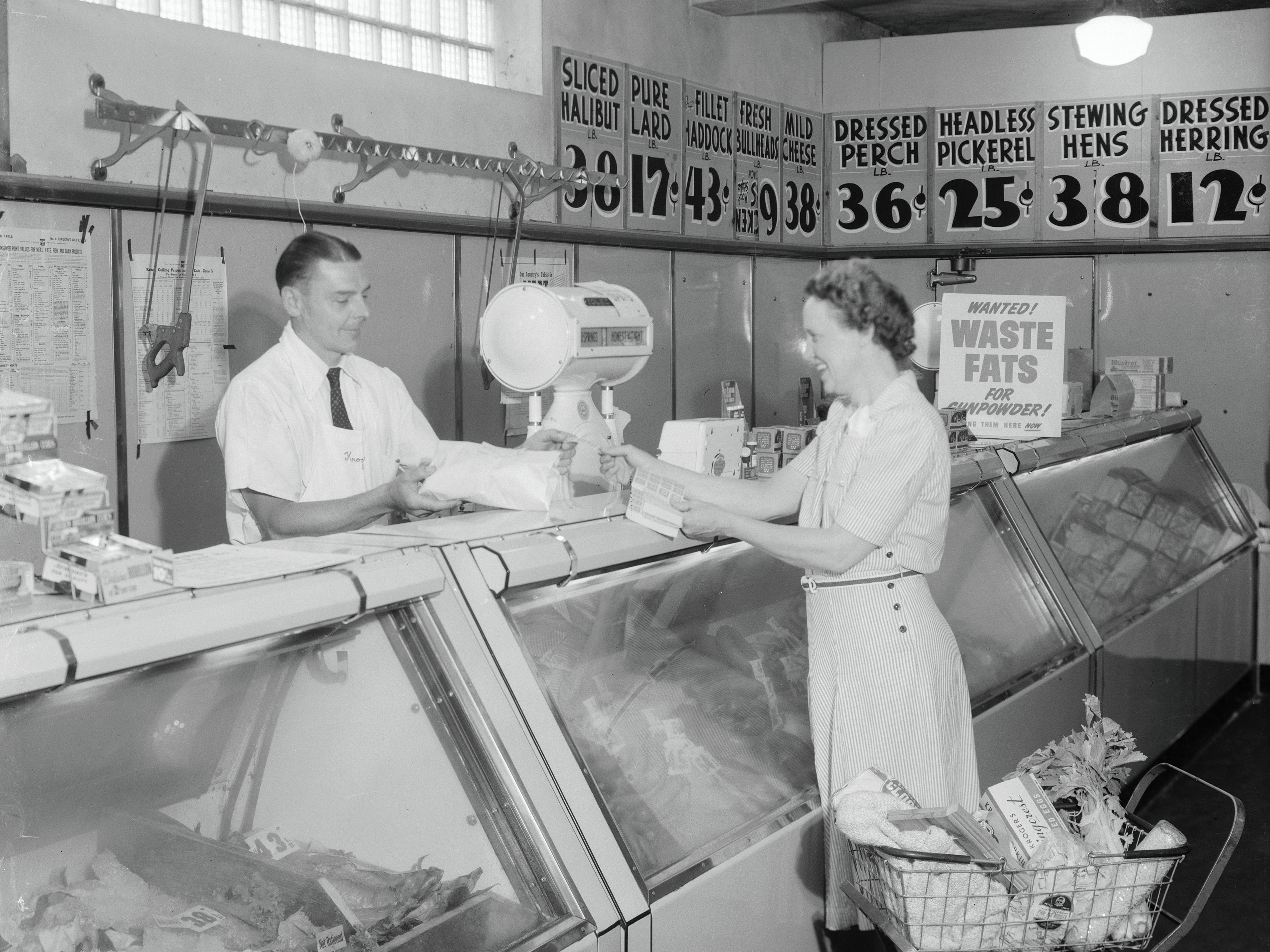 A woman stands next to a shopping cart while a butcher helps her at the meat counter