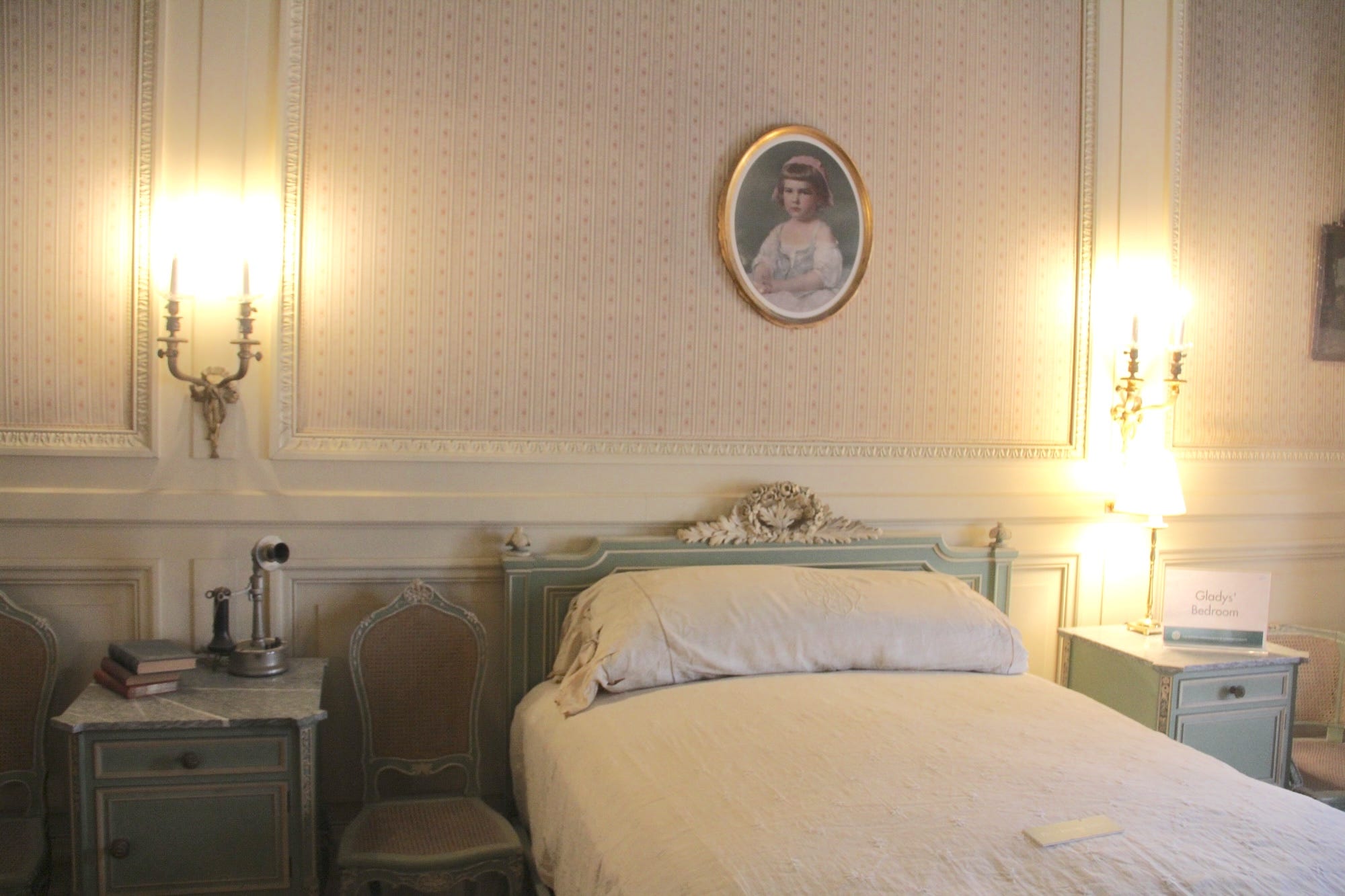 A bedroom in The Breakers with white wallpaper and a woman's portrait above the bed.