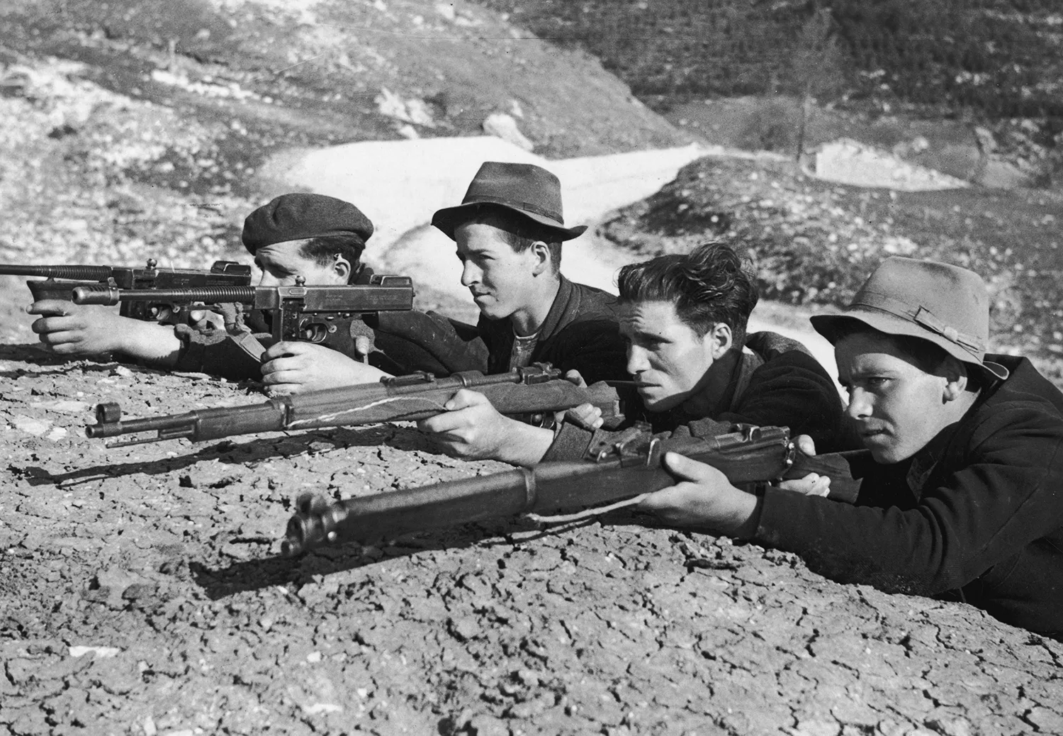 Four men squat down with rifles resting on the dirt in front of them.