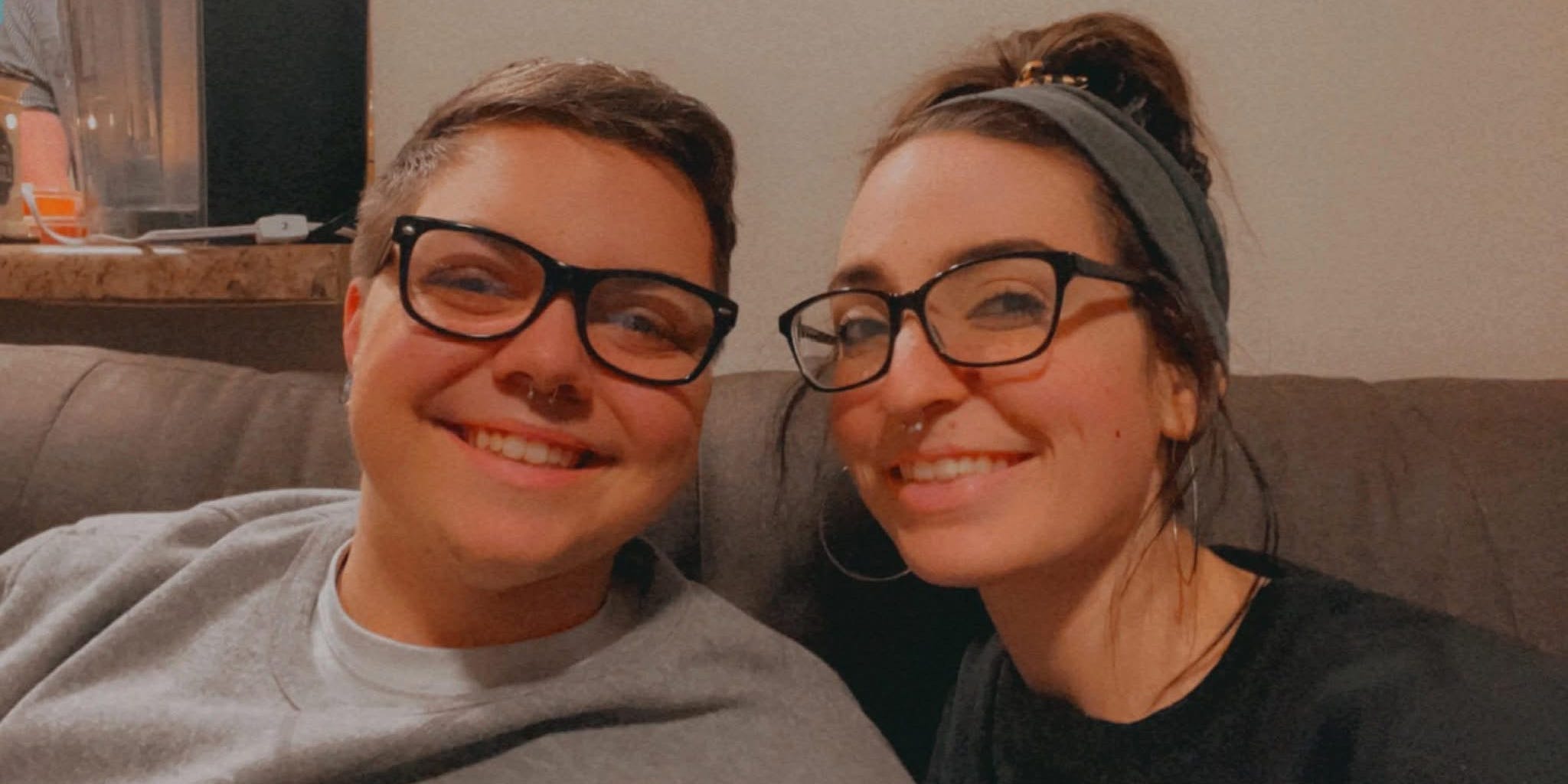In a selfie, a married queer couple smile while sitting on a couch.