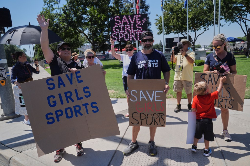 Spectators hold up signs protesting transgender athletes competing against biological females in high school sports.
