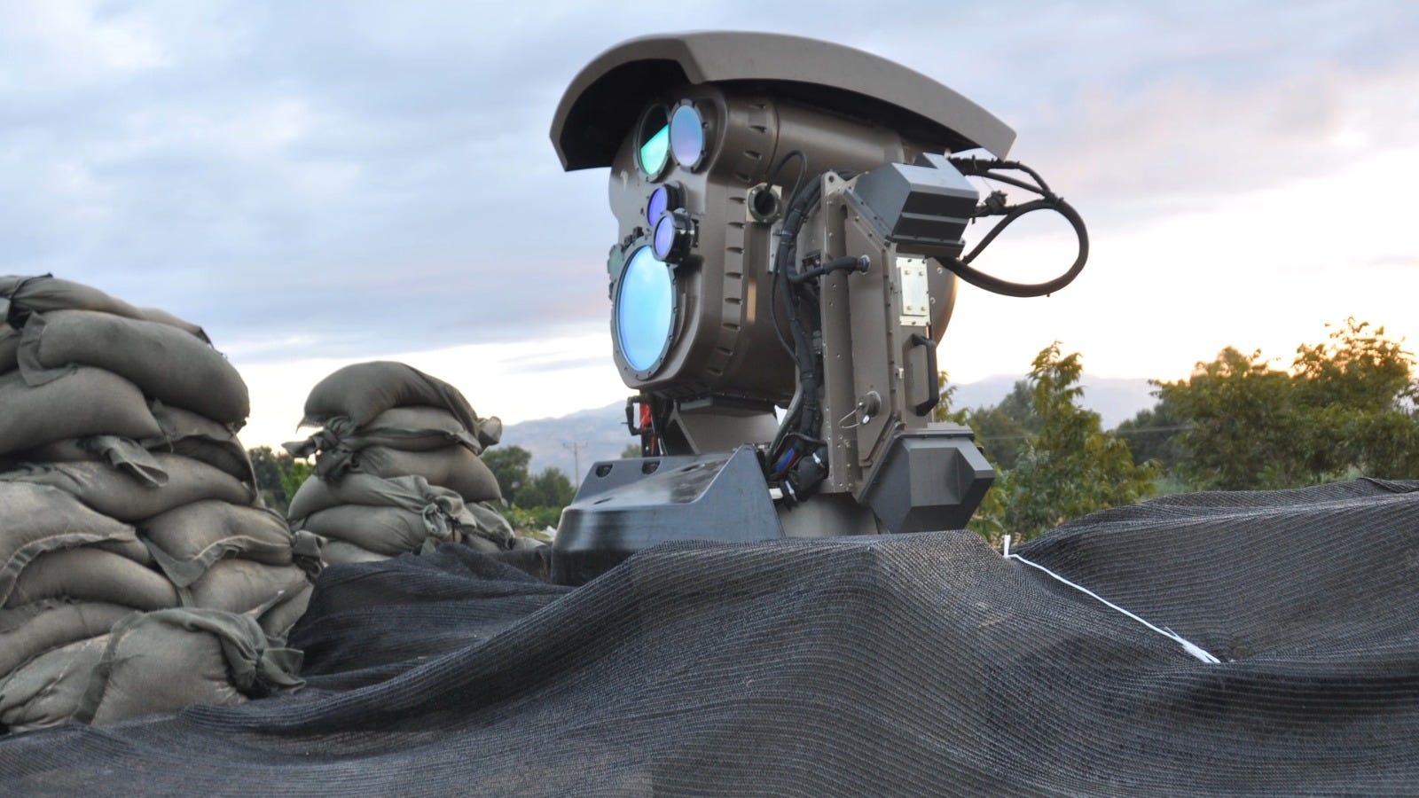 A side view of a laser weapon sitting on a tarp with sandbags nearby. The sky in the background is light and overcast.
