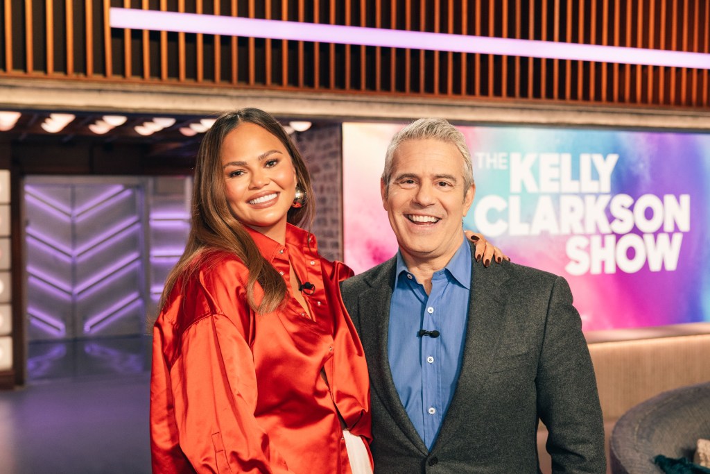 Chrissy Teigen in an orange satin shirt, hugs Andy Cohen in a charcoal grey suit and blue shirt.