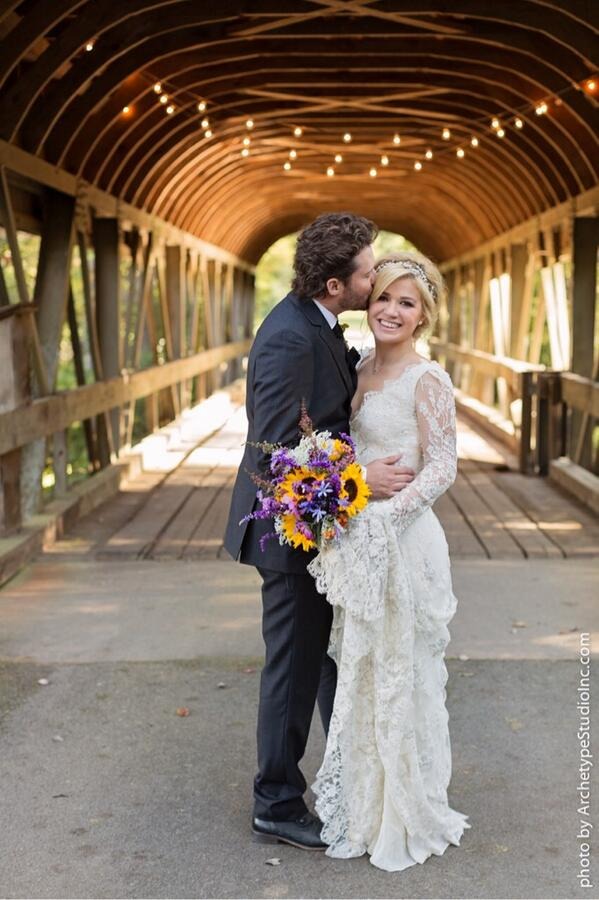 Kelly Clarkson in a lace wedding gown, holding a bouquet of sunflowers. Brandon Blackstock, wearing a dark suit, kisses her.
