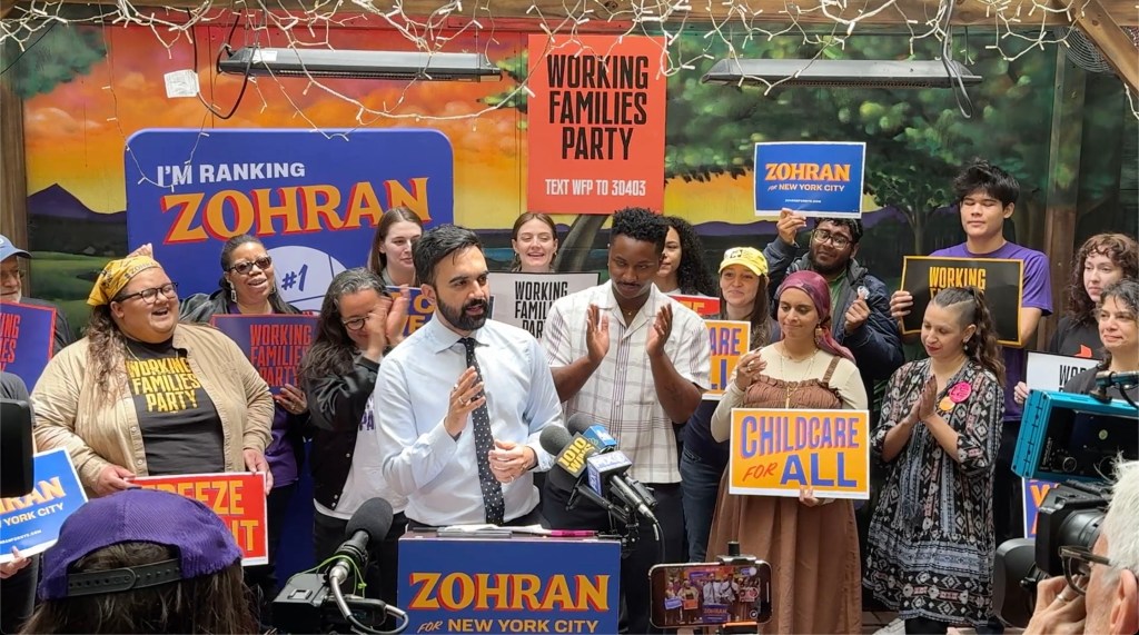 Zohran Mamdani speaking at a rally with working family party members and signs reading i'm ranking zohran and childcare for all 
