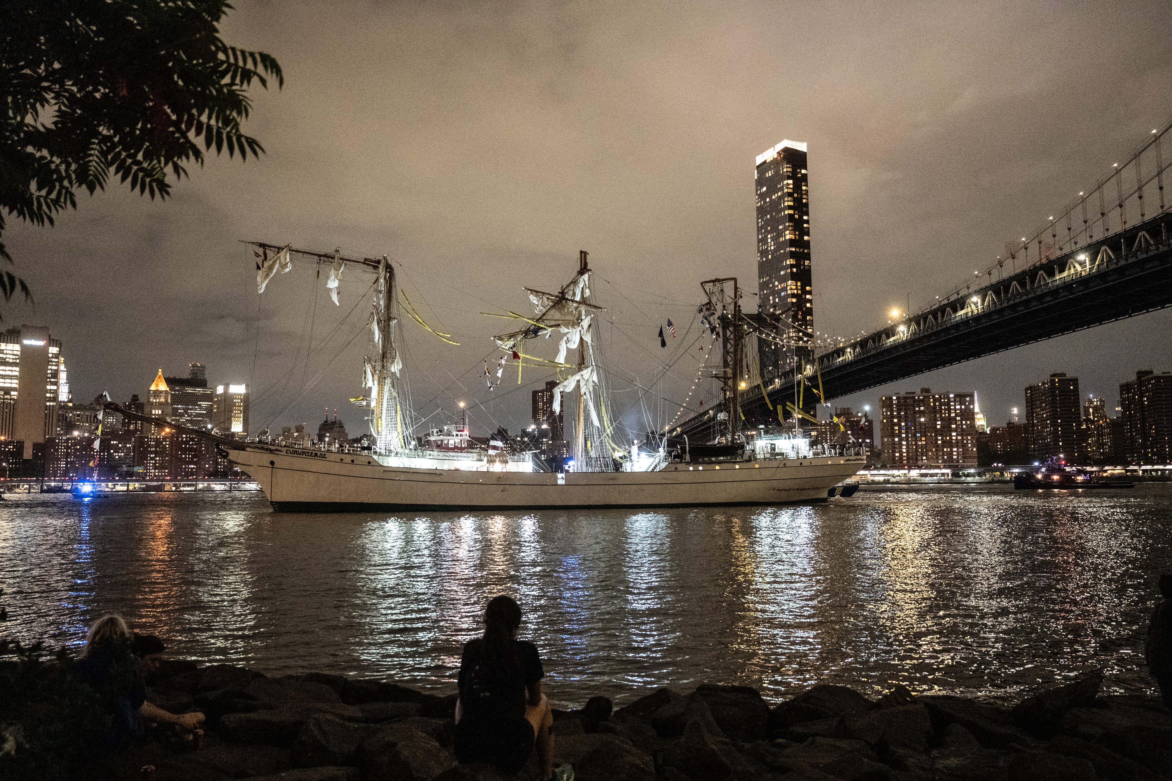 A masted ship with the tops of its masts destroyed in the East River by Brooklyn bridge