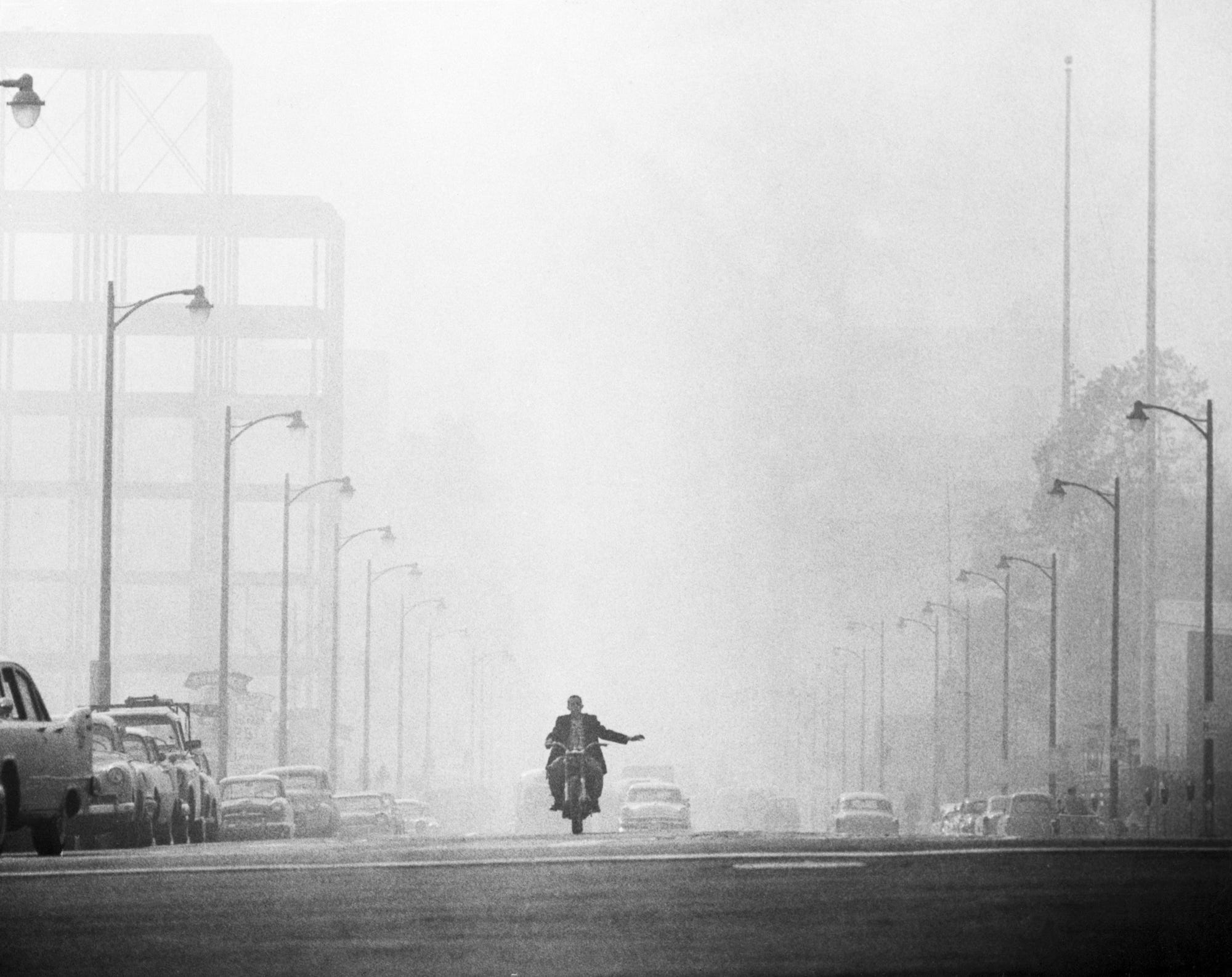 A motorcyclist in Los Angeles prepares to turn while driving along a street which is engulfed in a thick haze combined by fog and smog in 1958.