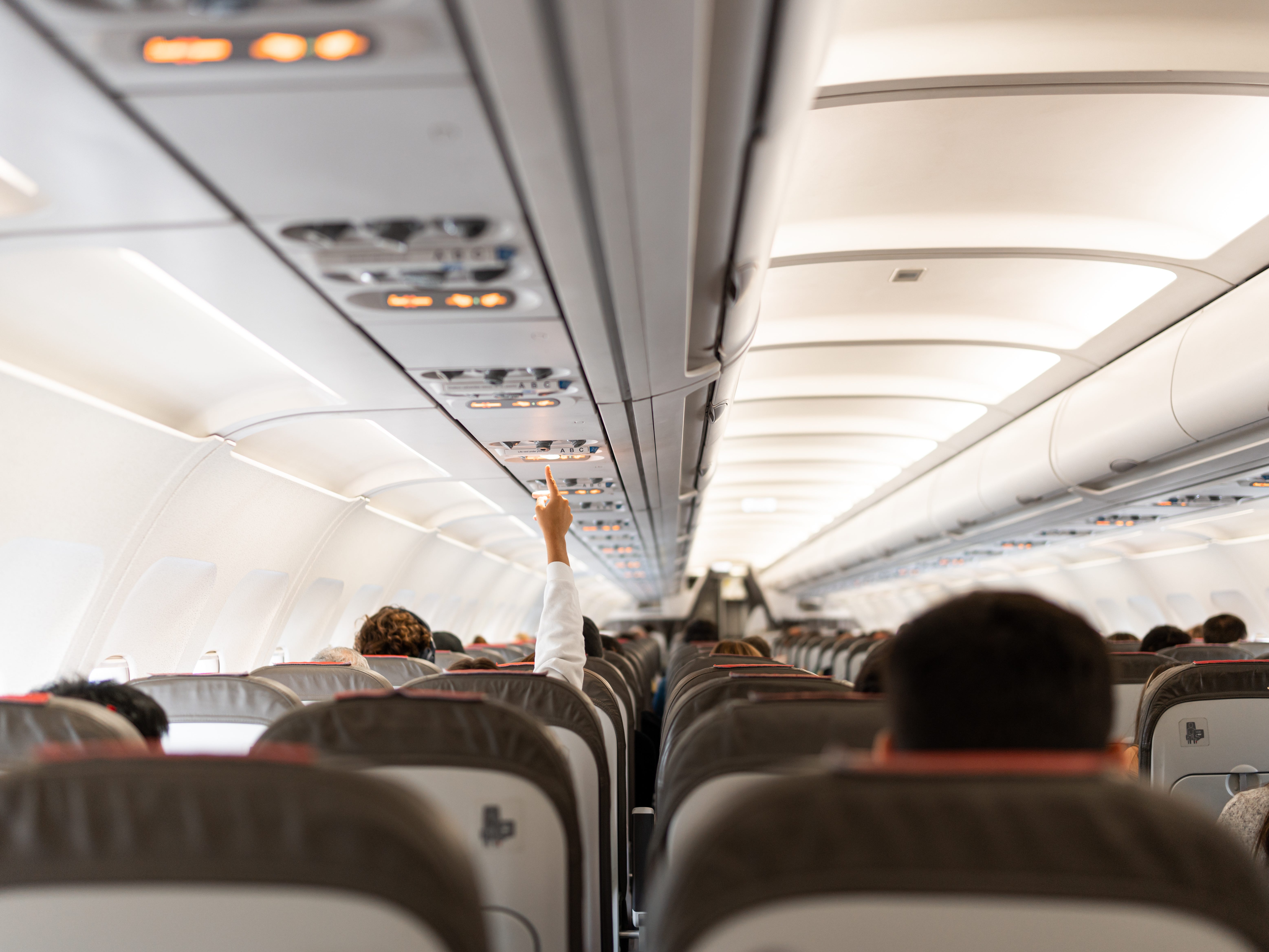 People sitting on rows of seats on a plane, with one person reaching their hand up to call for a flight attendant.