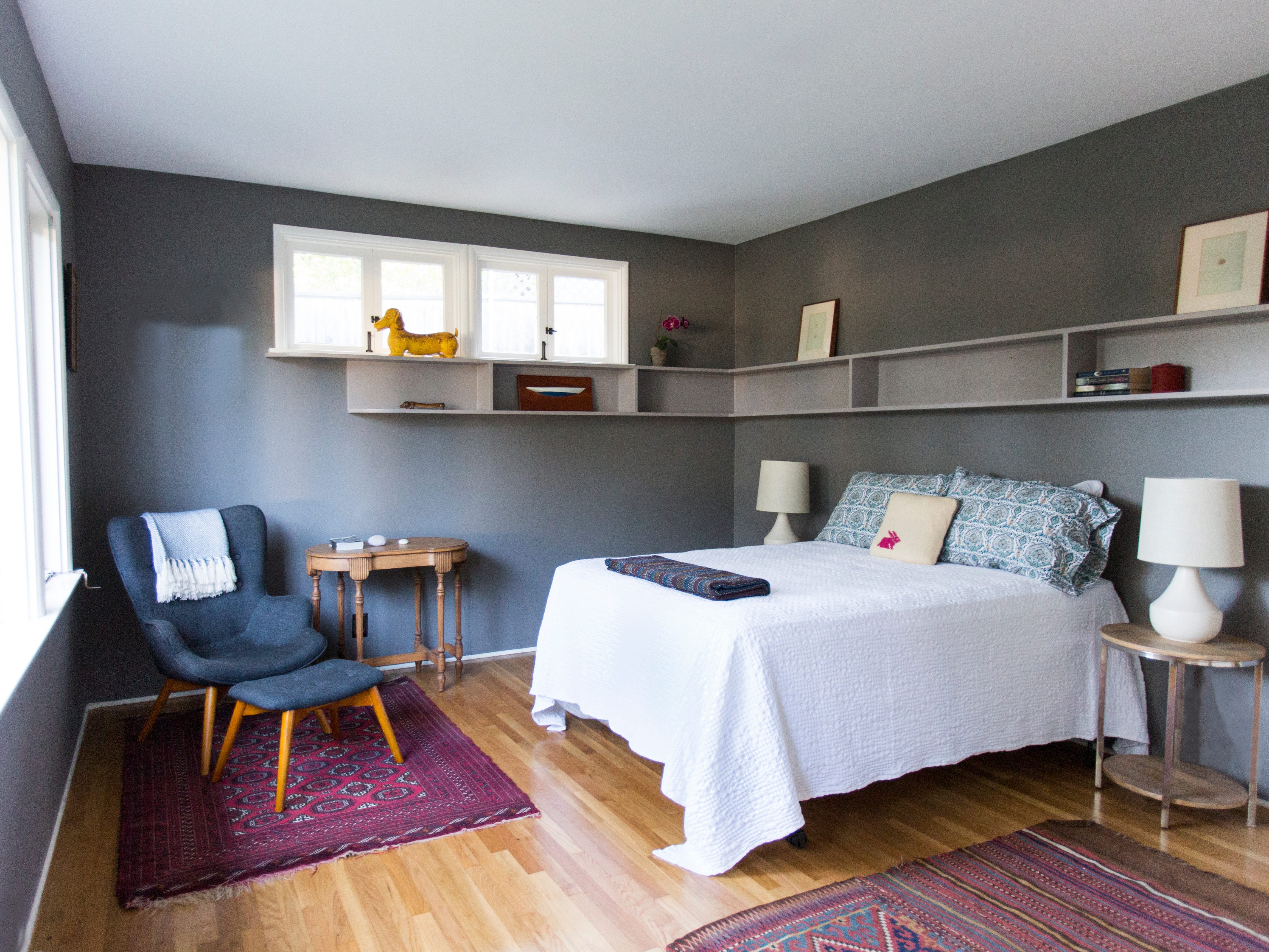 A bedroom with colorful rugs, a white and blue bedspread, a blue chair, a nightstand with a white lamp, and a gray shelf along the gray walls.