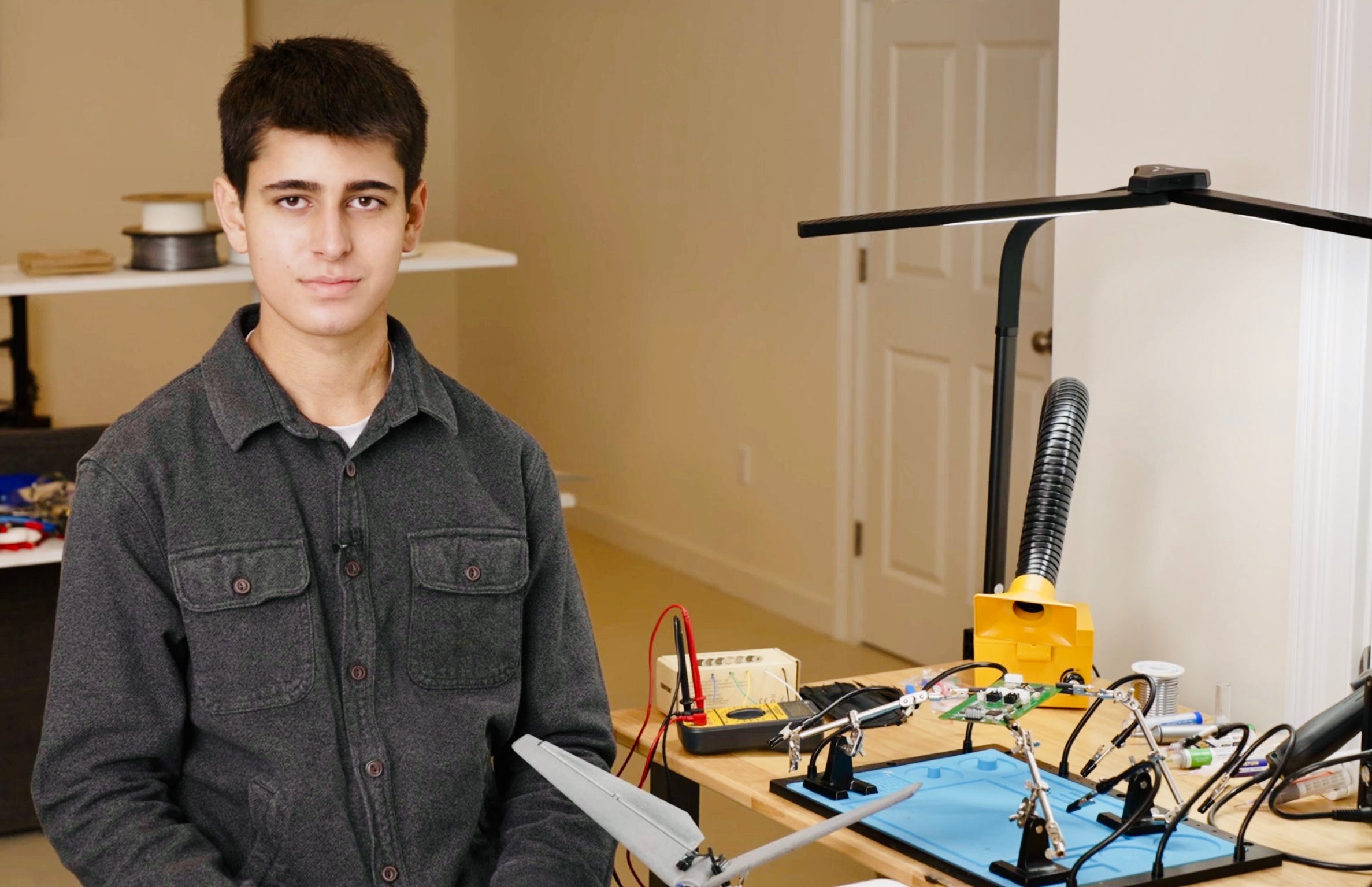 teenage boy sitting at a desk covered in electronics and wiring in a white room