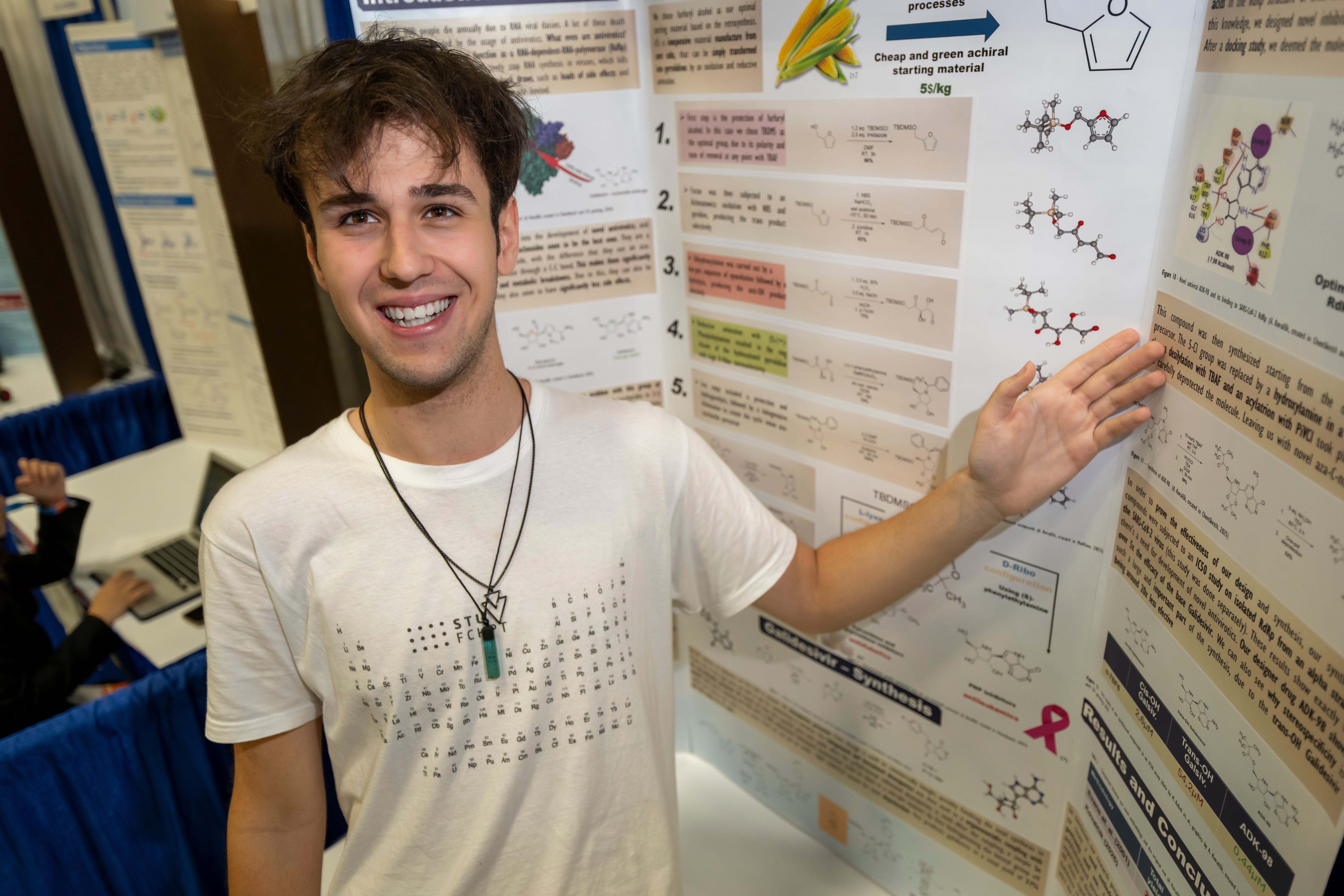 smiling young man in a white shirt gesturing at a scientific poster board with chemistry graphics