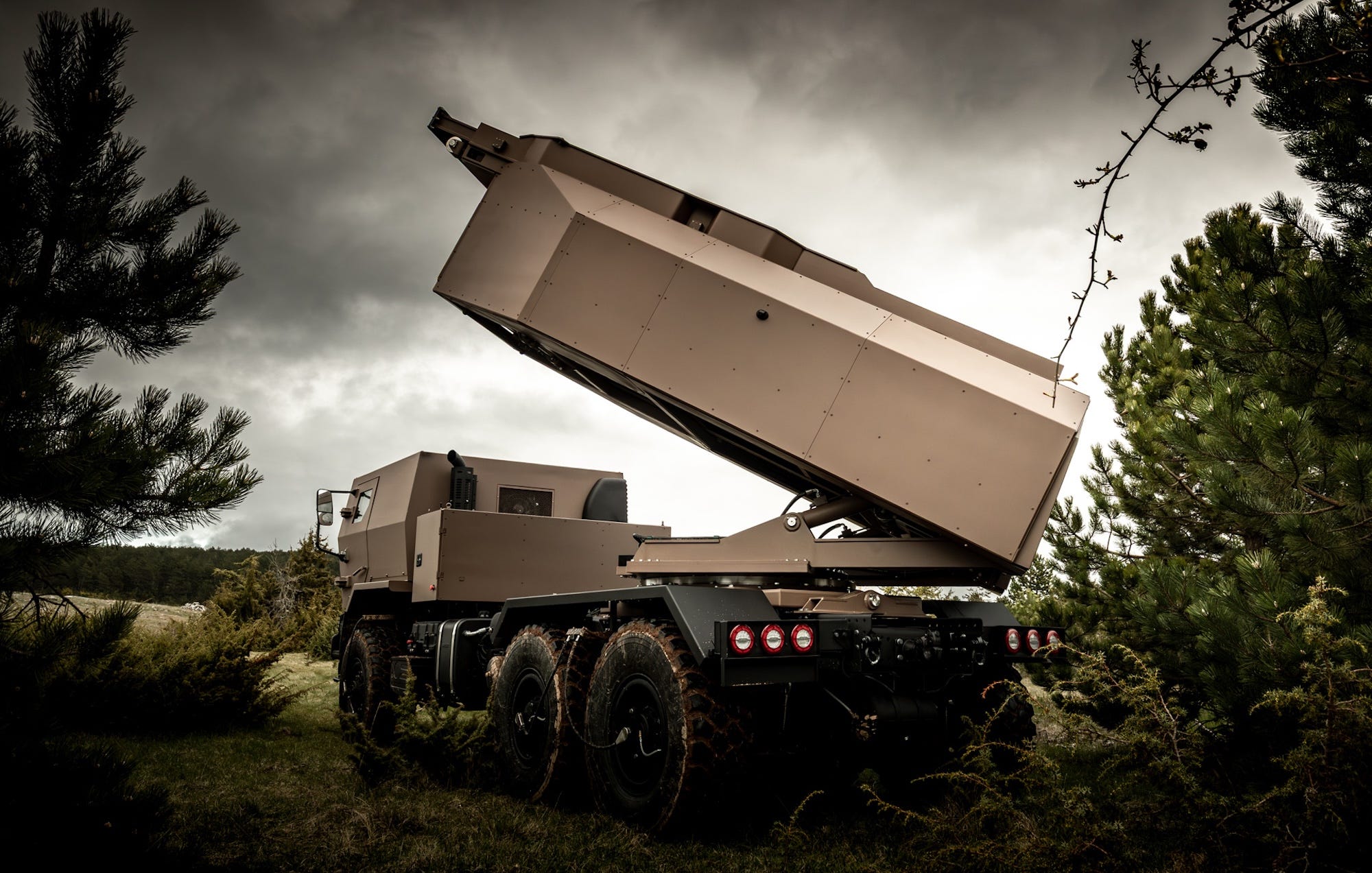 A light brown weapoins sytem mounted on a six-wheel truck between trees and in front of a cloudy sky
