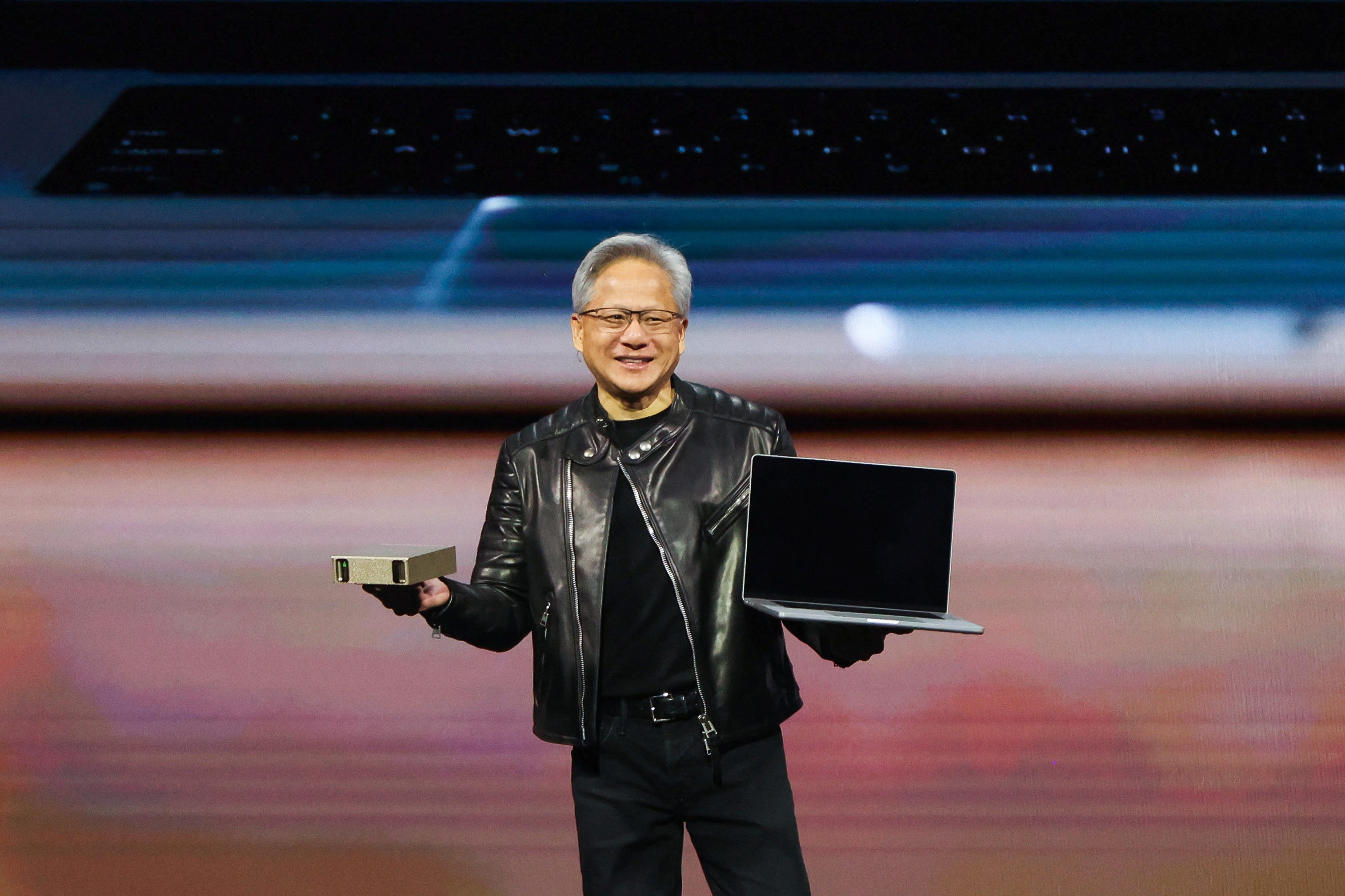 A man wearing all black and a leather jacket holds a consumer GPU and a laptop on a stage