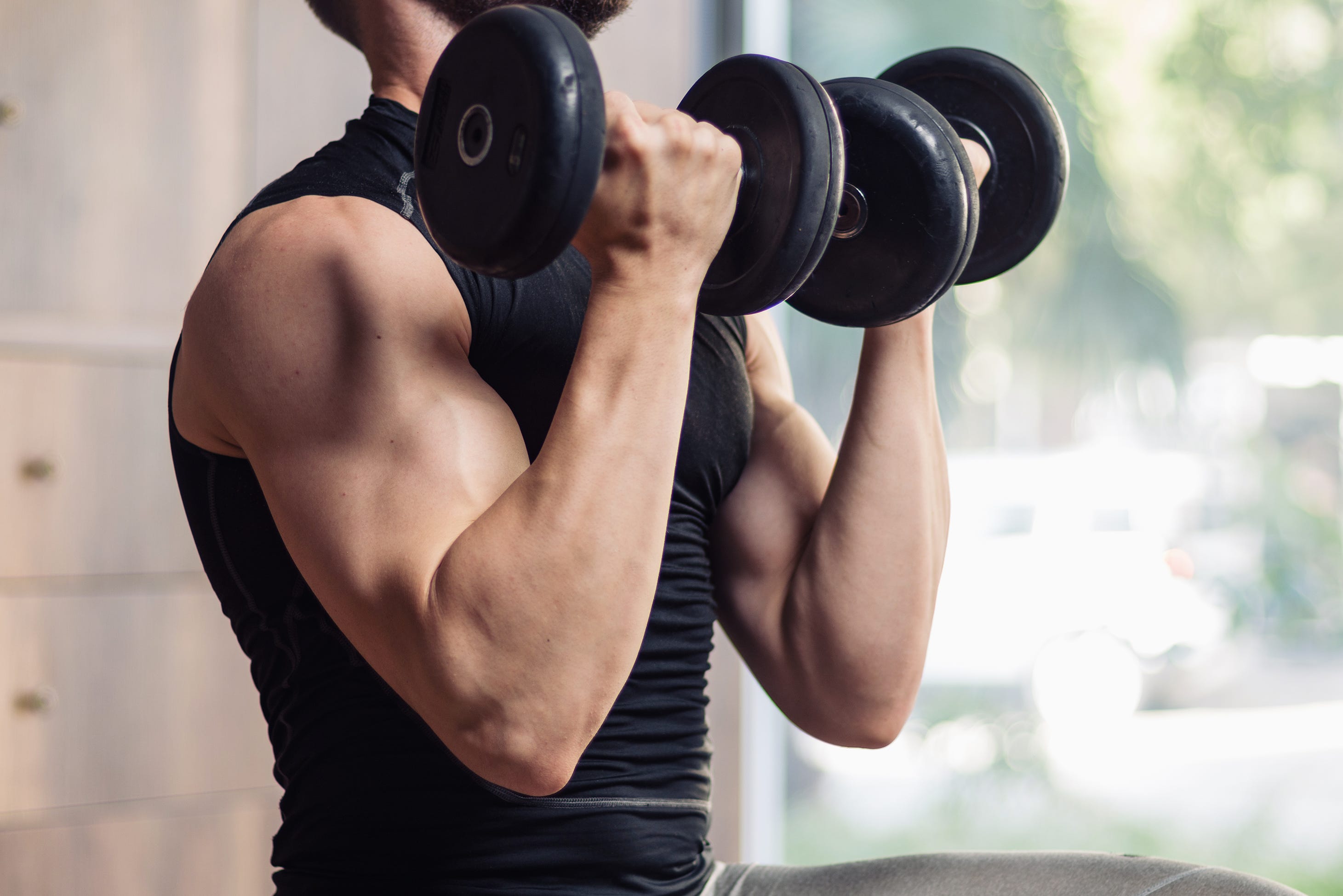 a close up of a man performing a dumbbell bicep curl in a gym in front of a sunny window