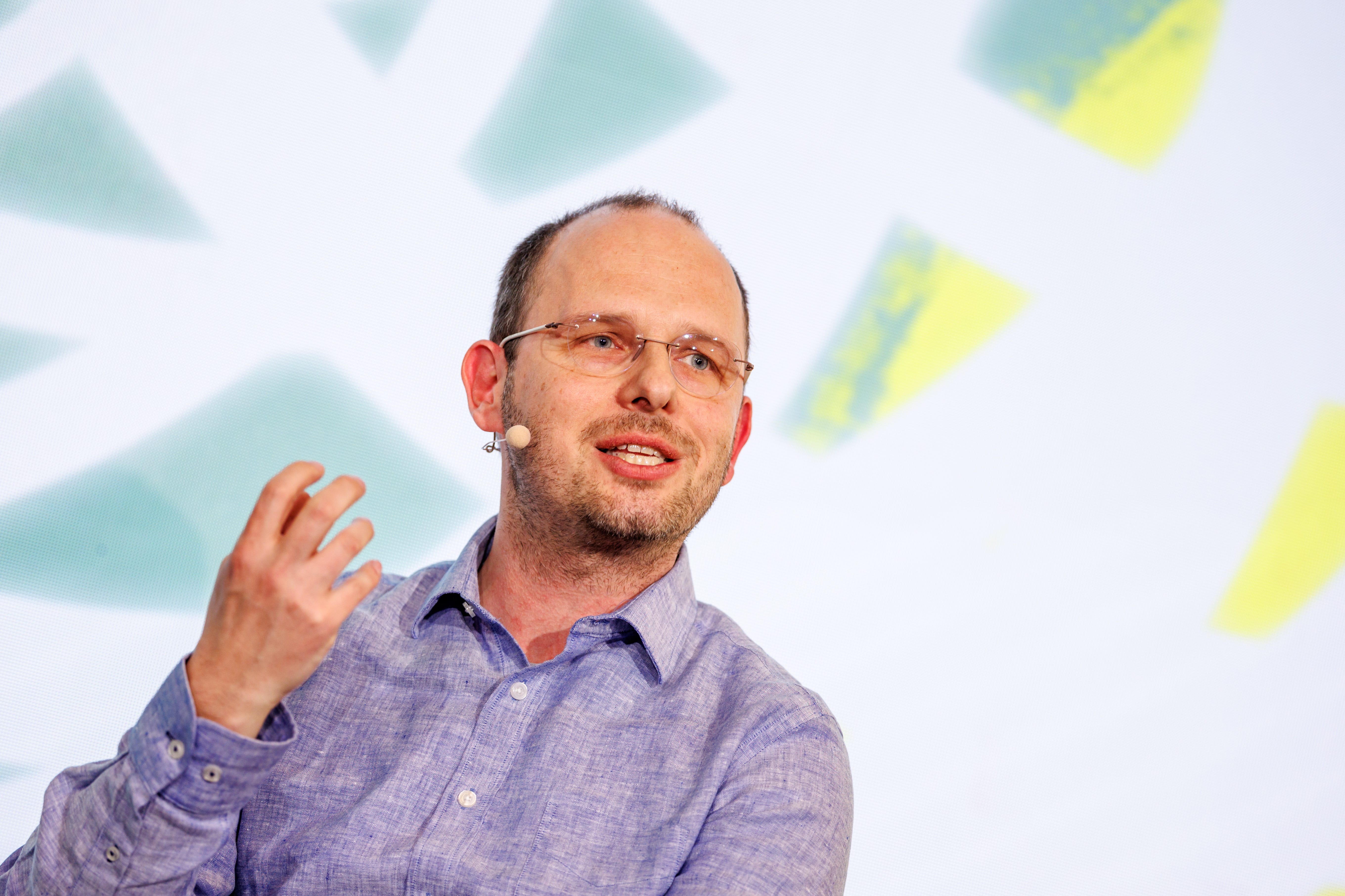 Thomas Dohmke, CEO of Github, wears glasses and a blue button down shirt, long-sleeved, in front of a blue-white background that is littered with blue and yellow geometric shapes. He is holding up his right hand, and is mid-speech.