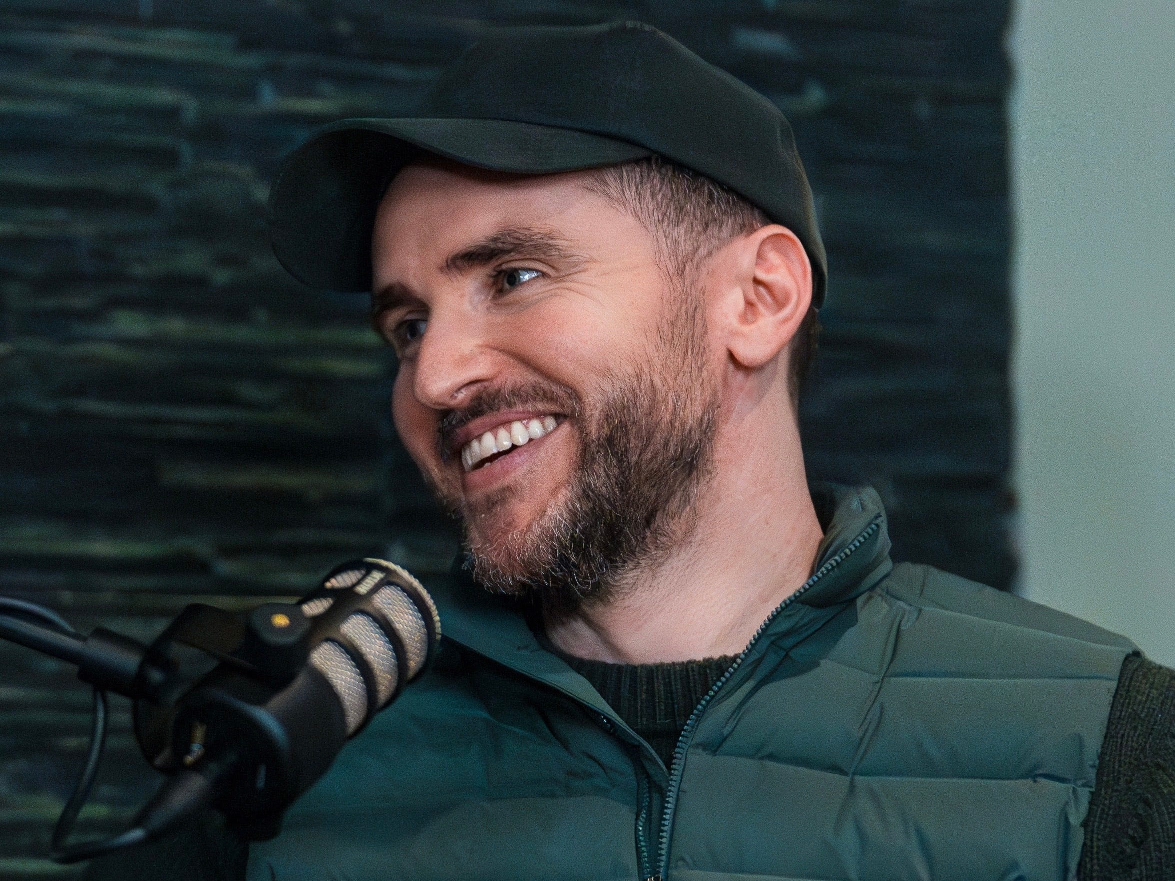 Tom Stokely smiling in front of a microphone, wearing a green puffer vest and a black baseball hat.