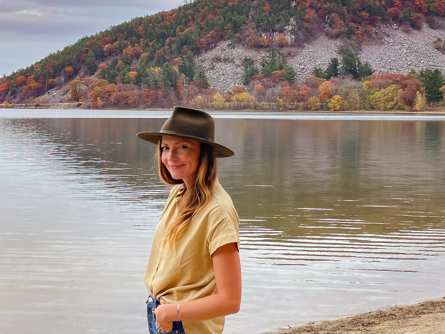 Emily stands in front of a lake with a mountain covered in fall-colored trees in the background.