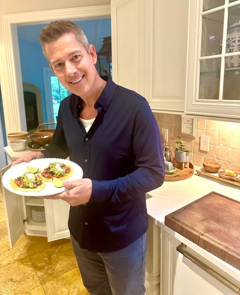 Sean Duffy in a kitchen in front of a white countertop and white wooden cabinets holding a plate with two tacos. He is wearing a dark blue untucked shirt with a low-cut t underneath and lighter casual trousers.