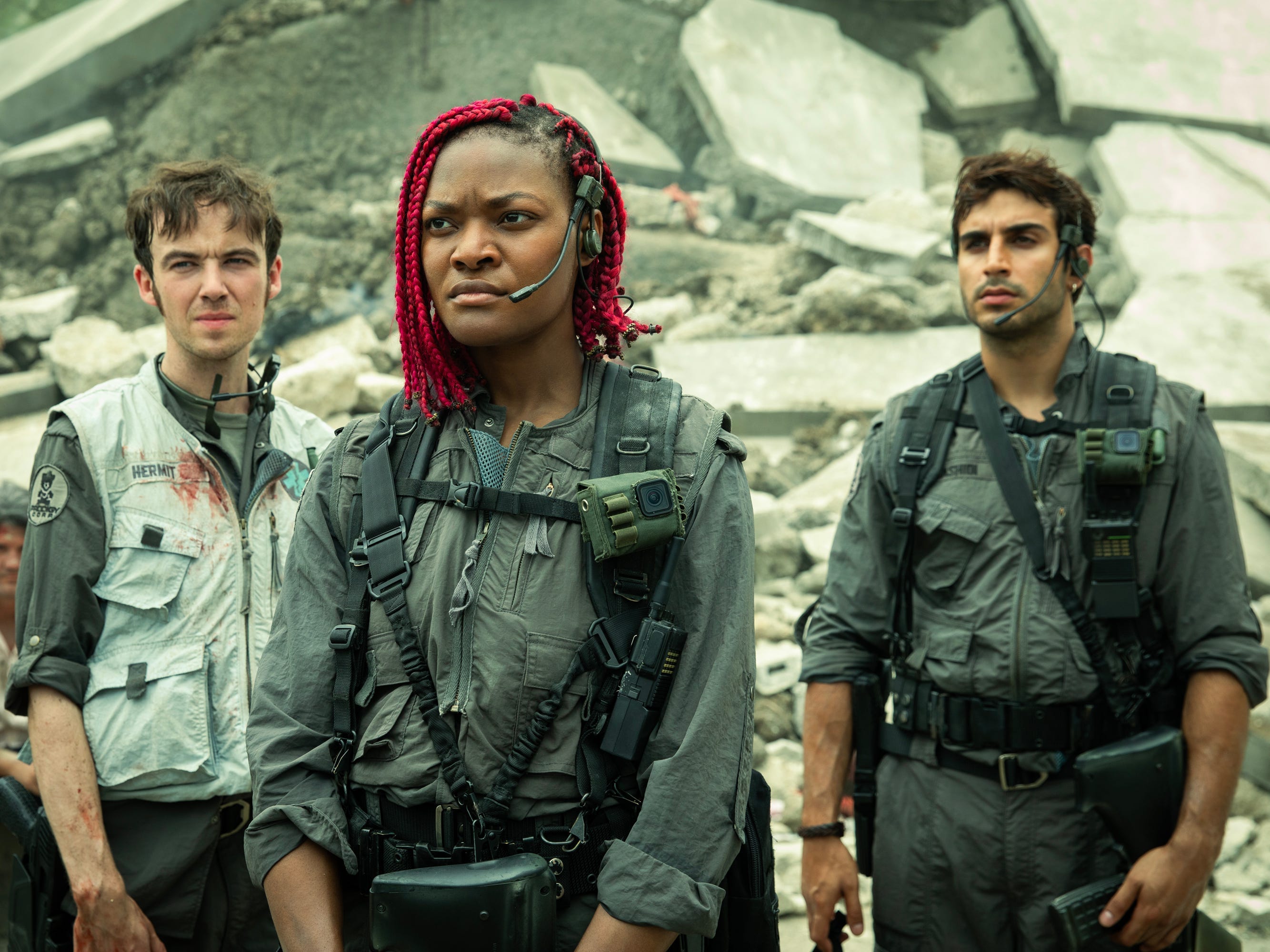 Three soldiers are standing in front of a pile of rubble. On the left is a man with brown hair wearing a blood-stained white vest over gray tactical gear. In the middle is a Black woman with red braids who is wearing gray and black tactical gear. ON the right is a man with brown hair and dark stubble. He's wearing the same tactical gear as the woman in the middle.