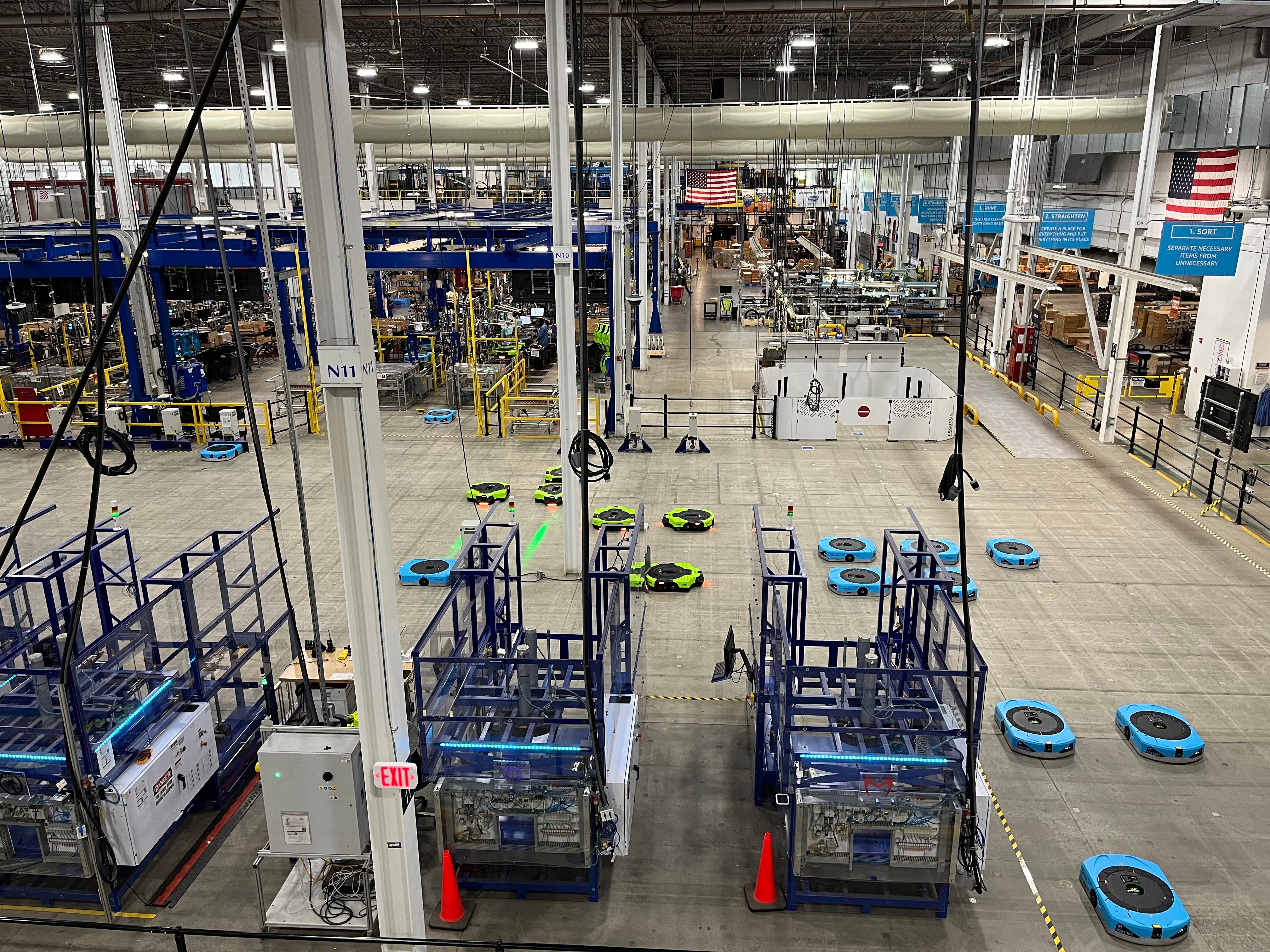 A view of an Amazon Robotics manufacturing facility shows blue and green mobile robots in the foreground and assembly lines in the background