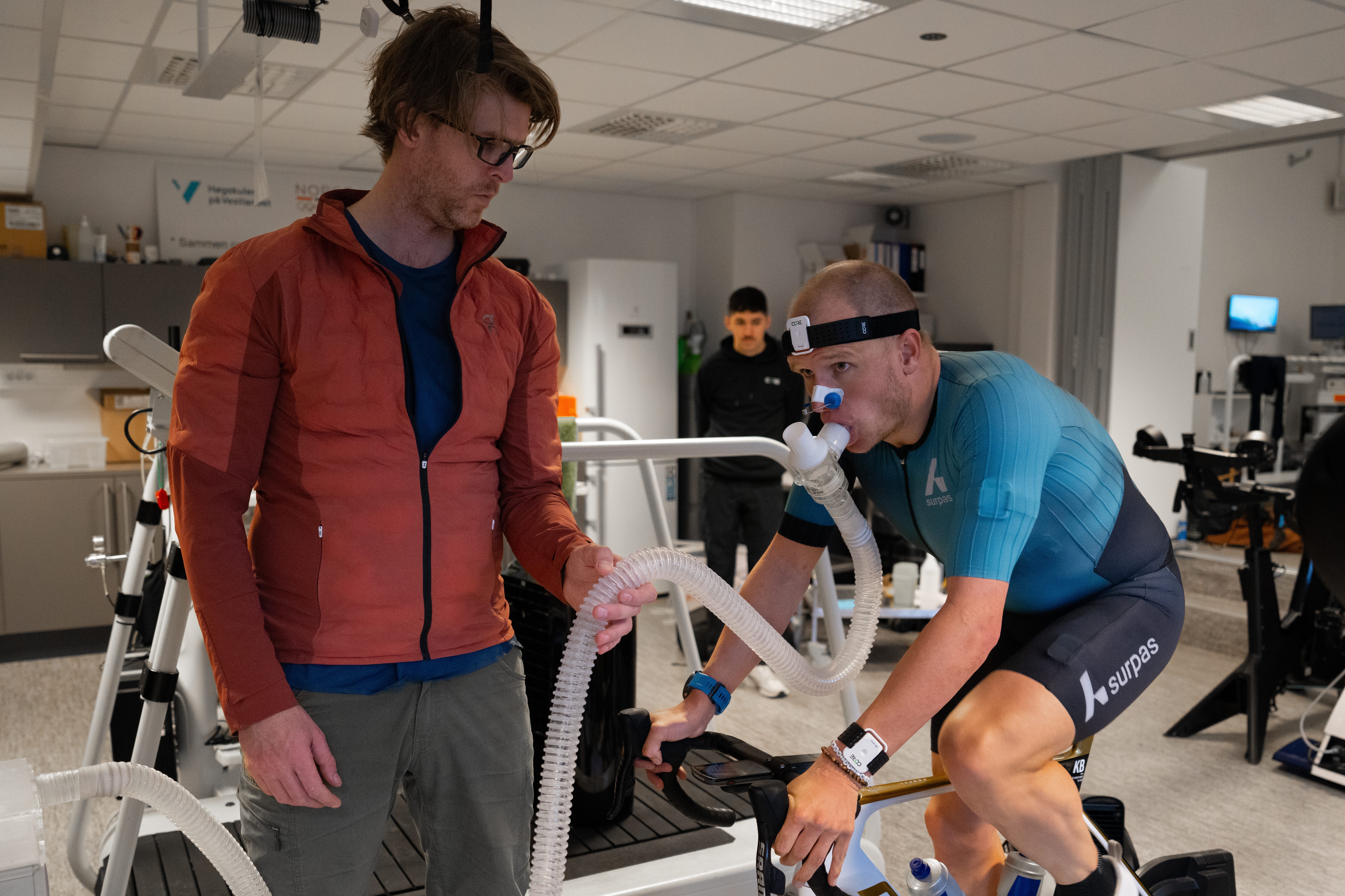A man on an exercise bike is performing an endurance test in a lab as his coach looks on.