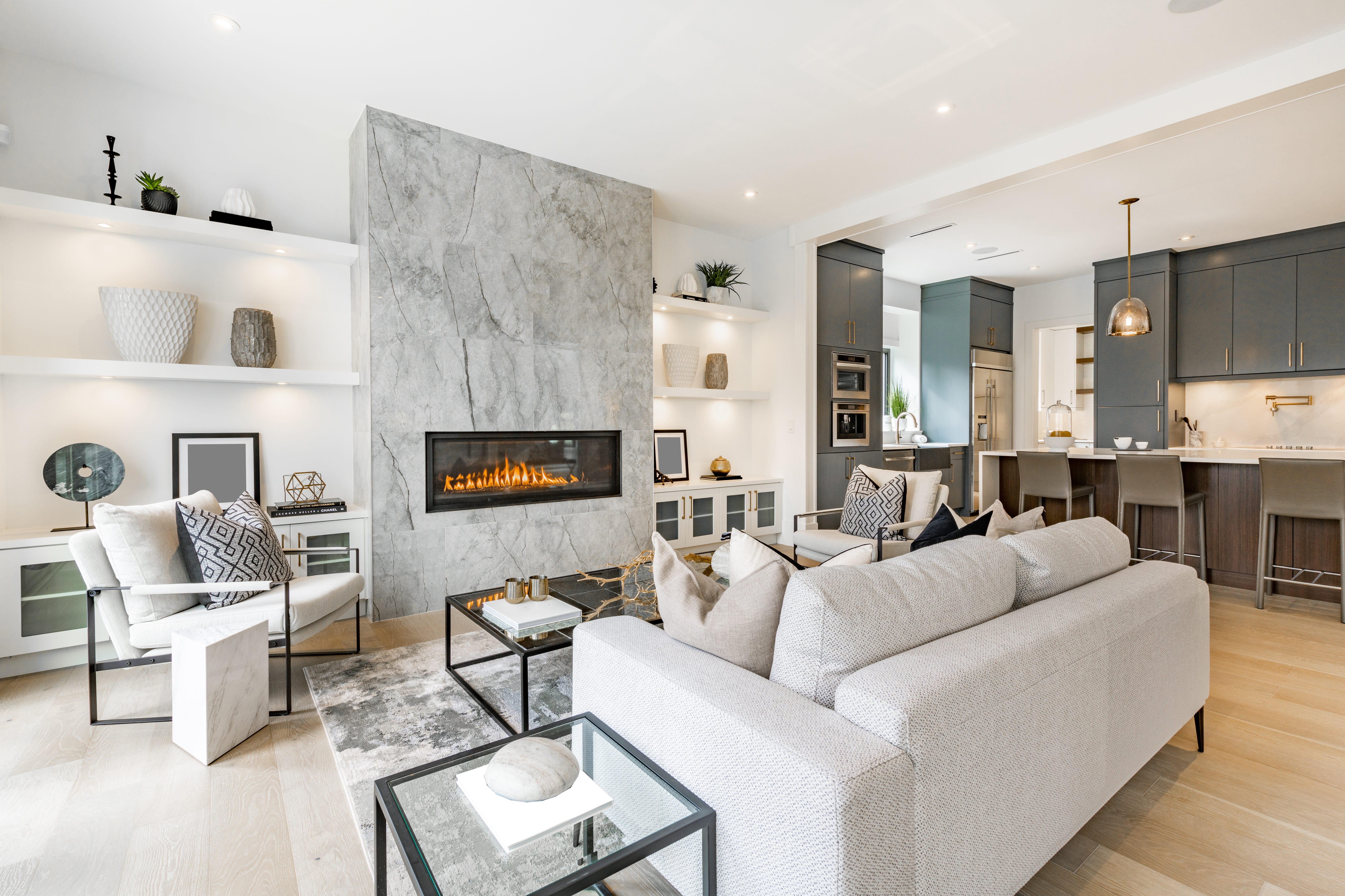 Living room with marble wall with faux fireplace open concept into kitchen with glass coffee tables and light-colored couches