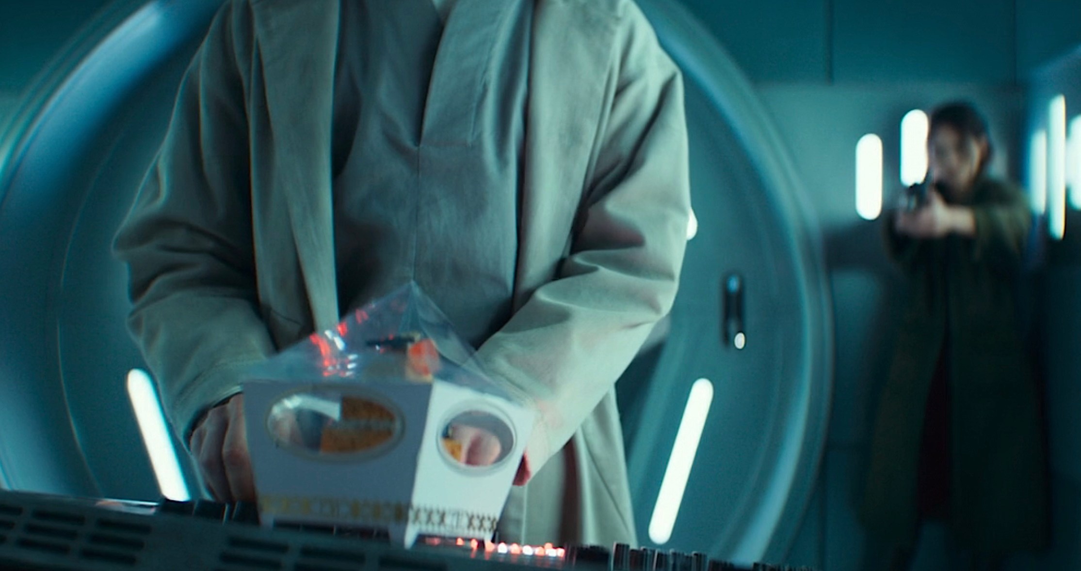 A shot from season 2, episode 6 of Andor, with Bix (Adria Arjona) in the background, out of focus, pointing a blaster at Imperial torturer Doctor Gorst (Joshua James), whose back is to her. He’s standing in the foreground at a computer panel, having just put down a fancy cupcake in a decorative box on the panel in front of him, in the extreme foreground.