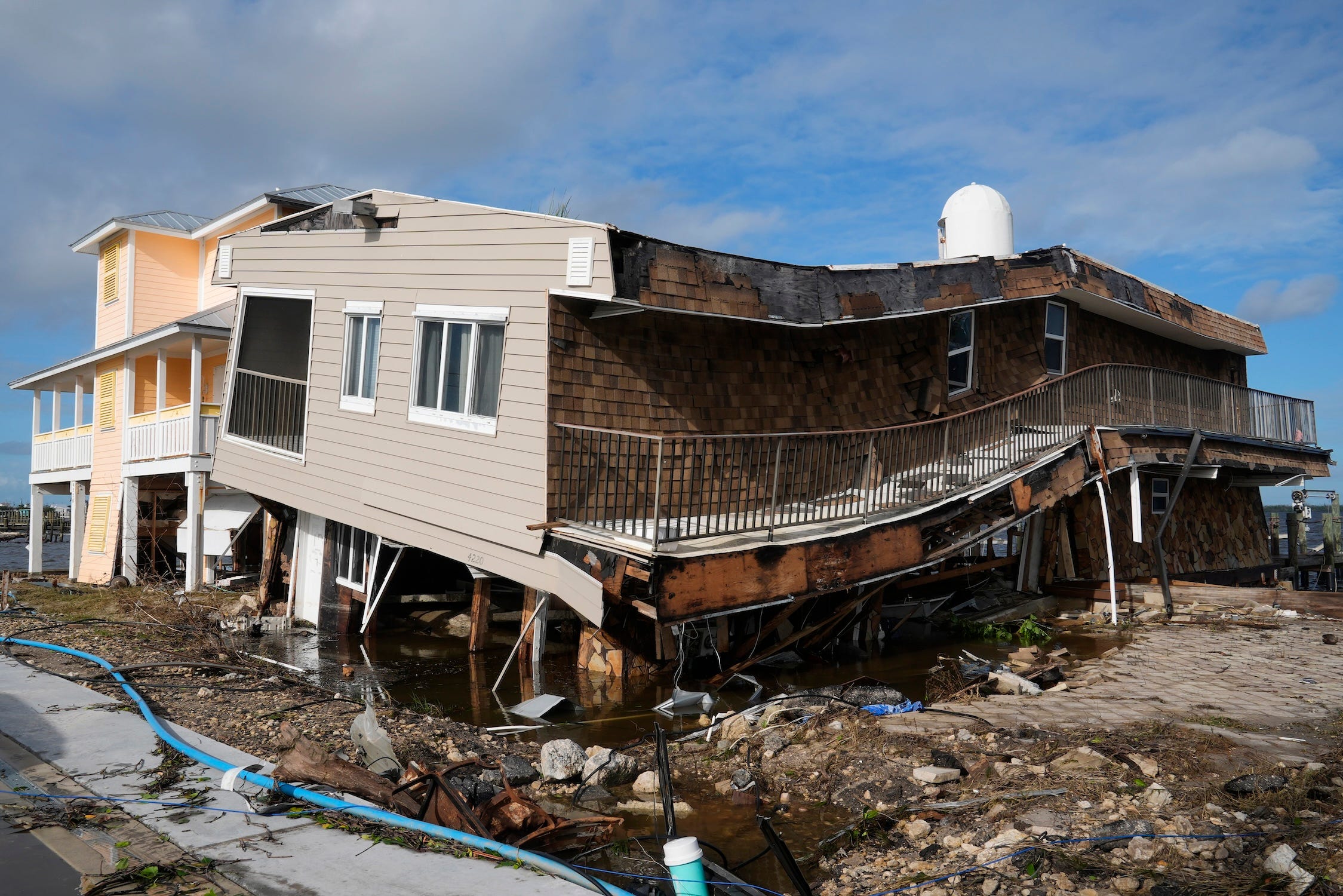 slumping one-story grey and wood panel house with panels ripped away next to a standing yellow house