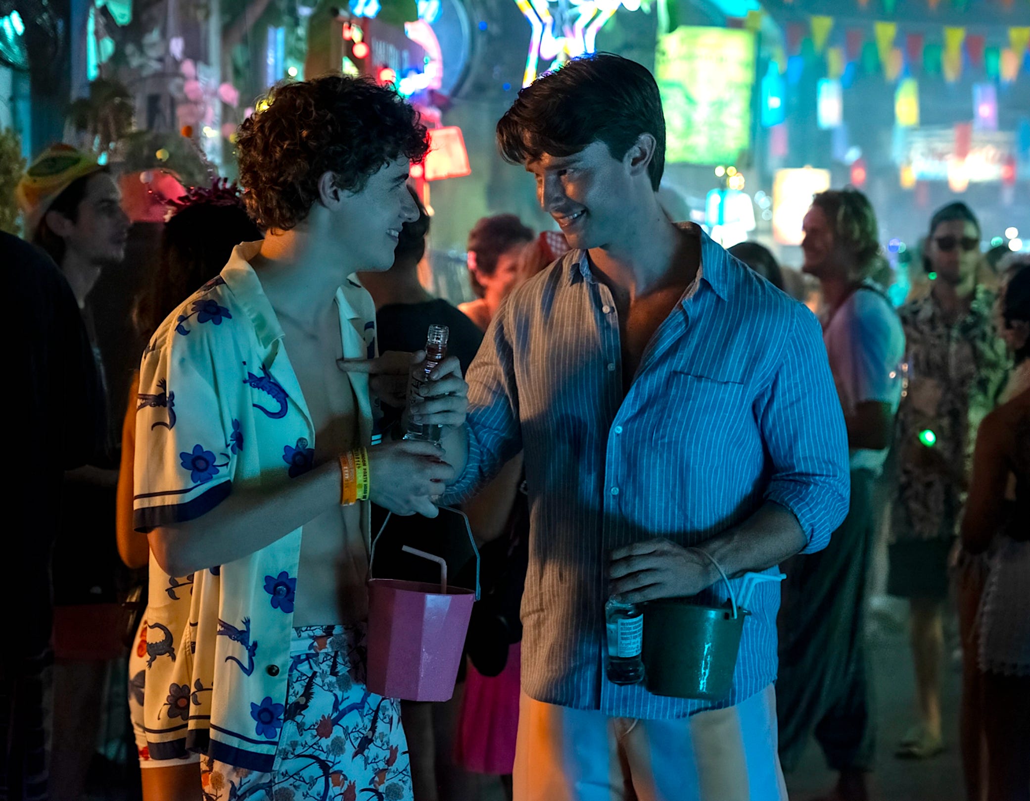 Two young men with brown hair are standing in a crowded street with neon lights and multicoloured flags behind them. On the left, a man is wearing an open white shirt with blue crocodiles and flowers printed on it. He's holding a bottle and a pink bucket with a straw in it in his left hand. On the right, the other man is wearing a striped blue shirt and is holding a green bucket and a bottle.