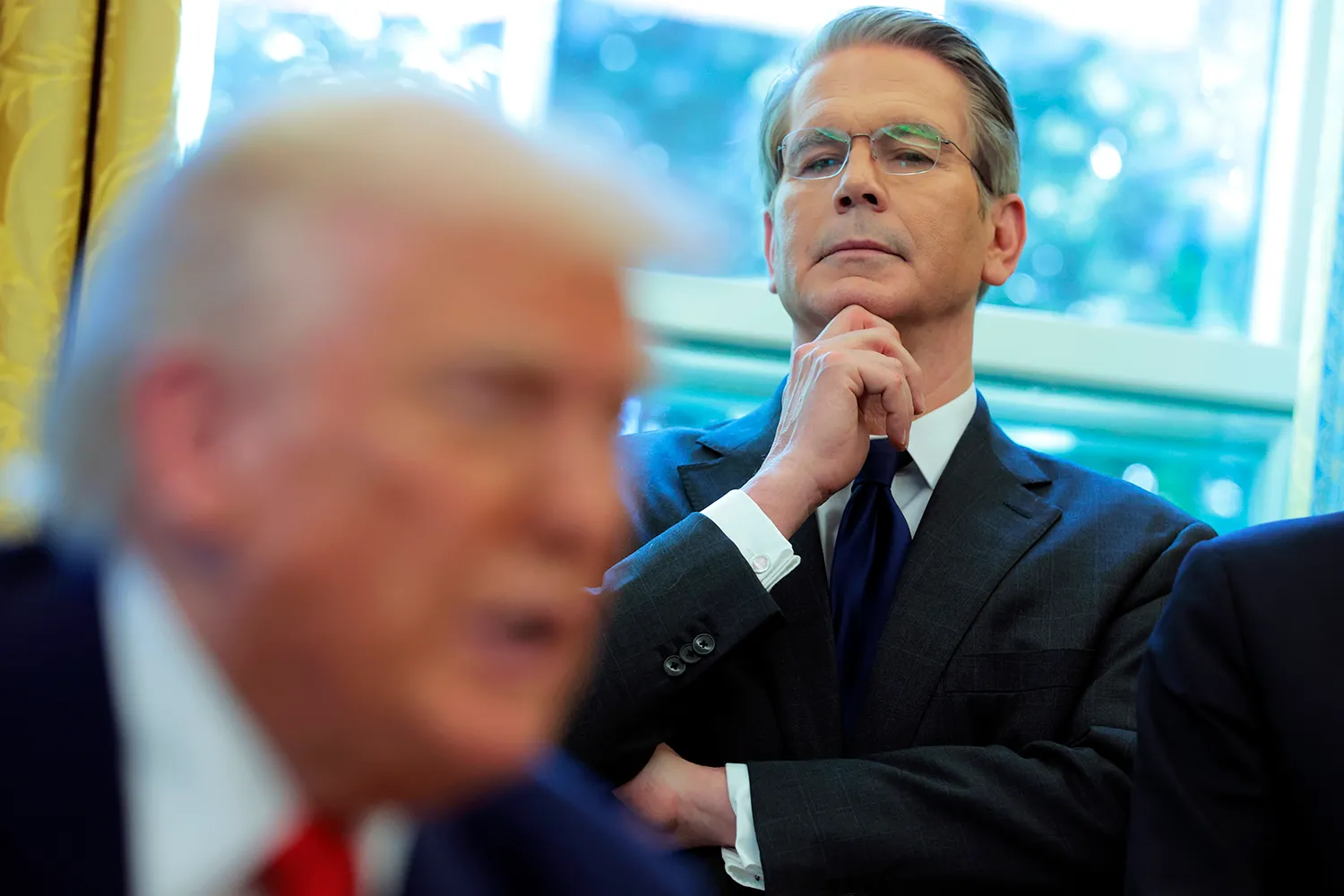 Scott Bessent, a man with short gray hair and glasses, rests his chin on his hand while watching Donald Trump, who is seen seated in the foreground, out of focus. Both men wear dark suits and ties.