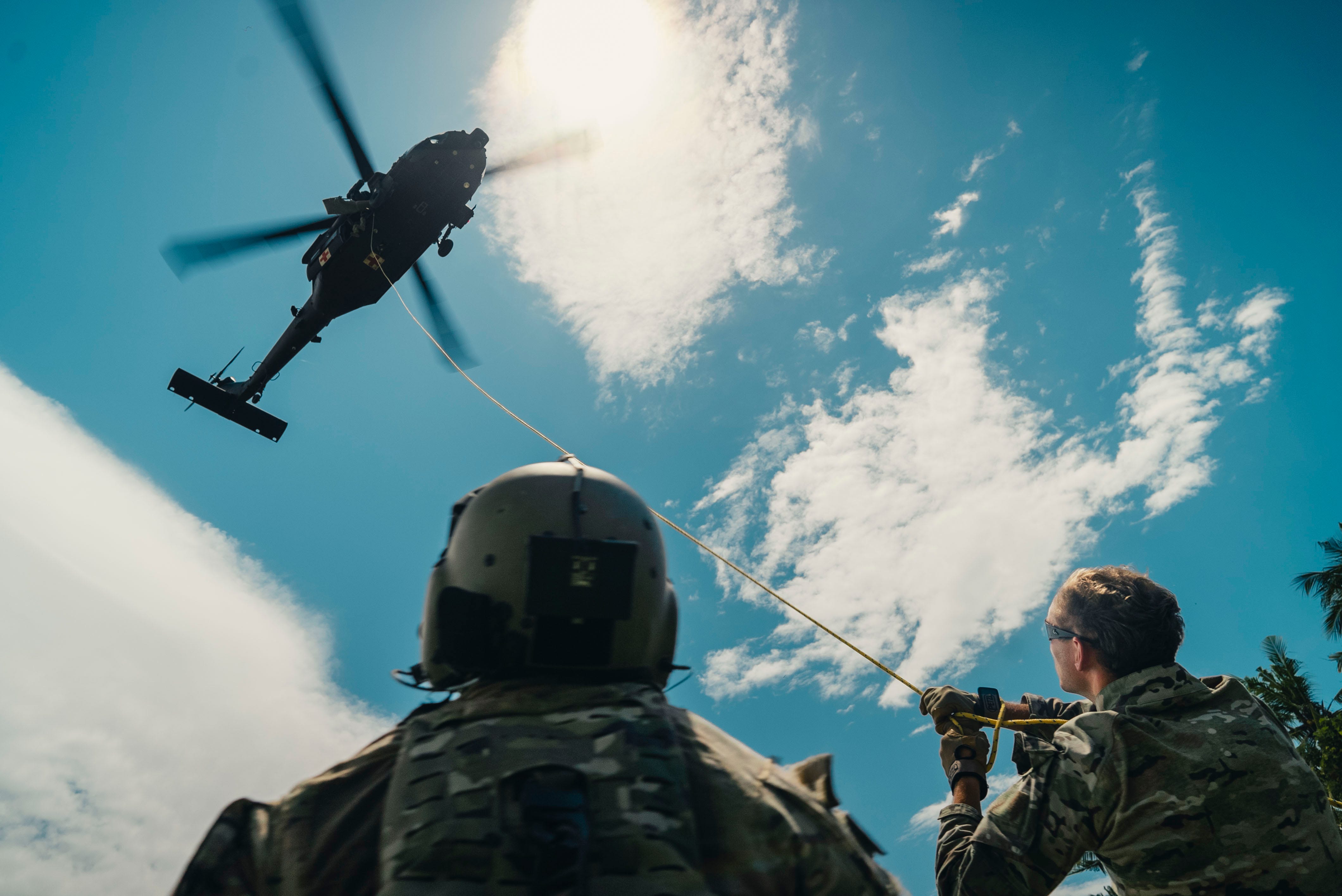 Two men wearing camouflage look up at a helicopter in a blue and cloudy sky.