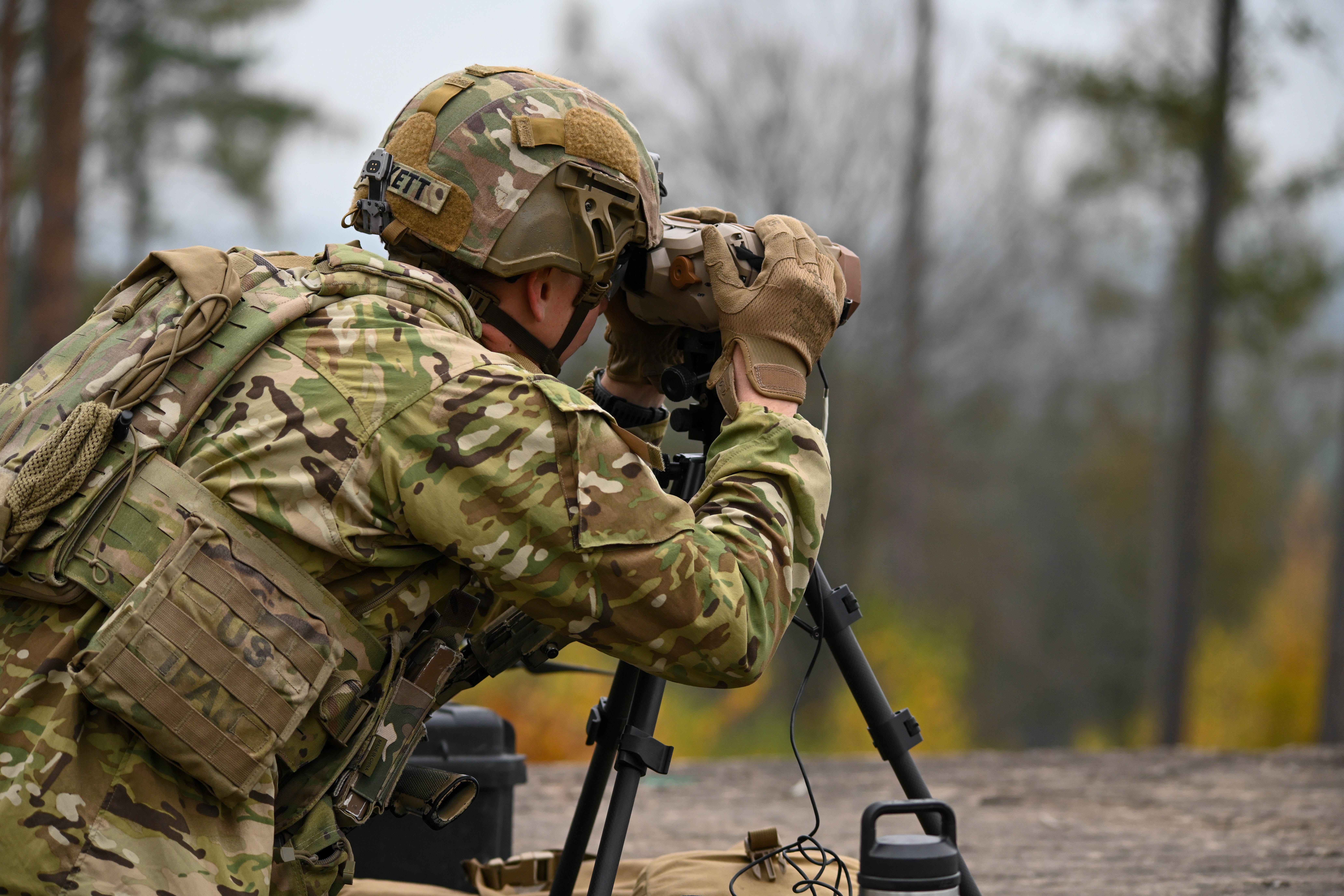 A soldier wearing camouflage looks through a scope on a tripod with blurred out trees and a grey sky in the background.