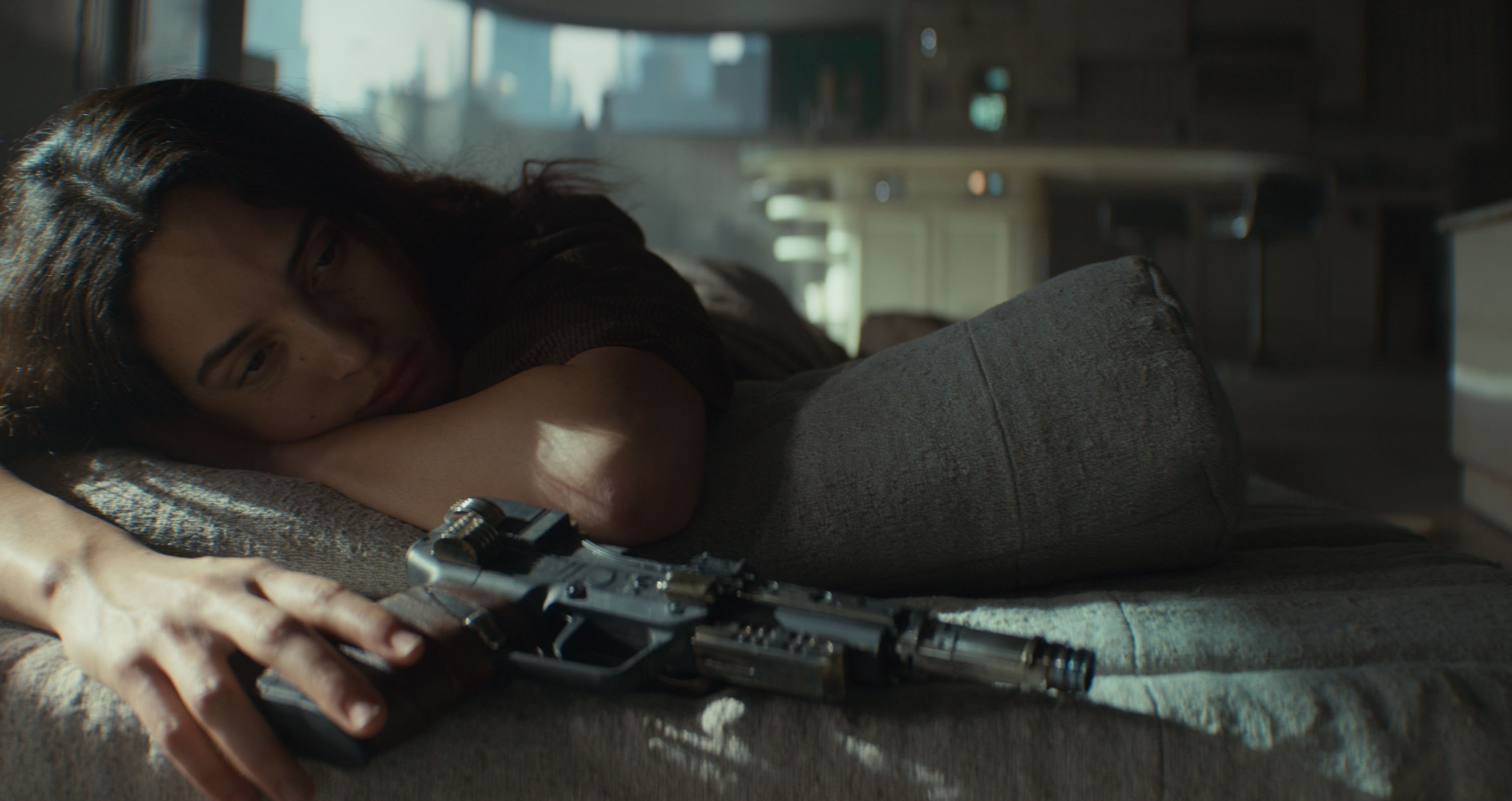 Bix Caleen (Adria Arjona) lies wearily on a bed in a dark room, fingers resting on the butt of a blaster, in Andor season 2