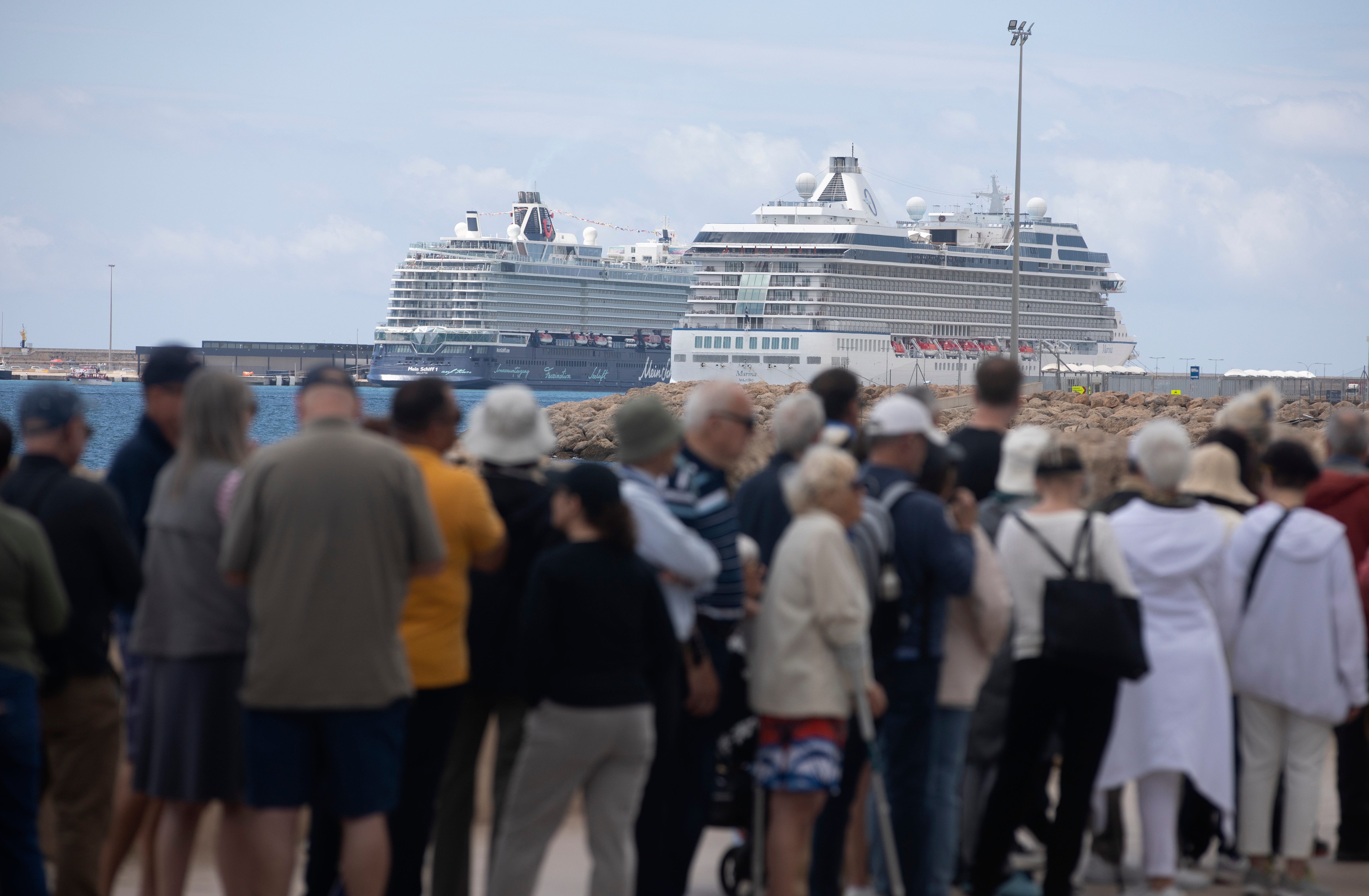 People wait to board 2 cruise ships anchored offshore in the background.