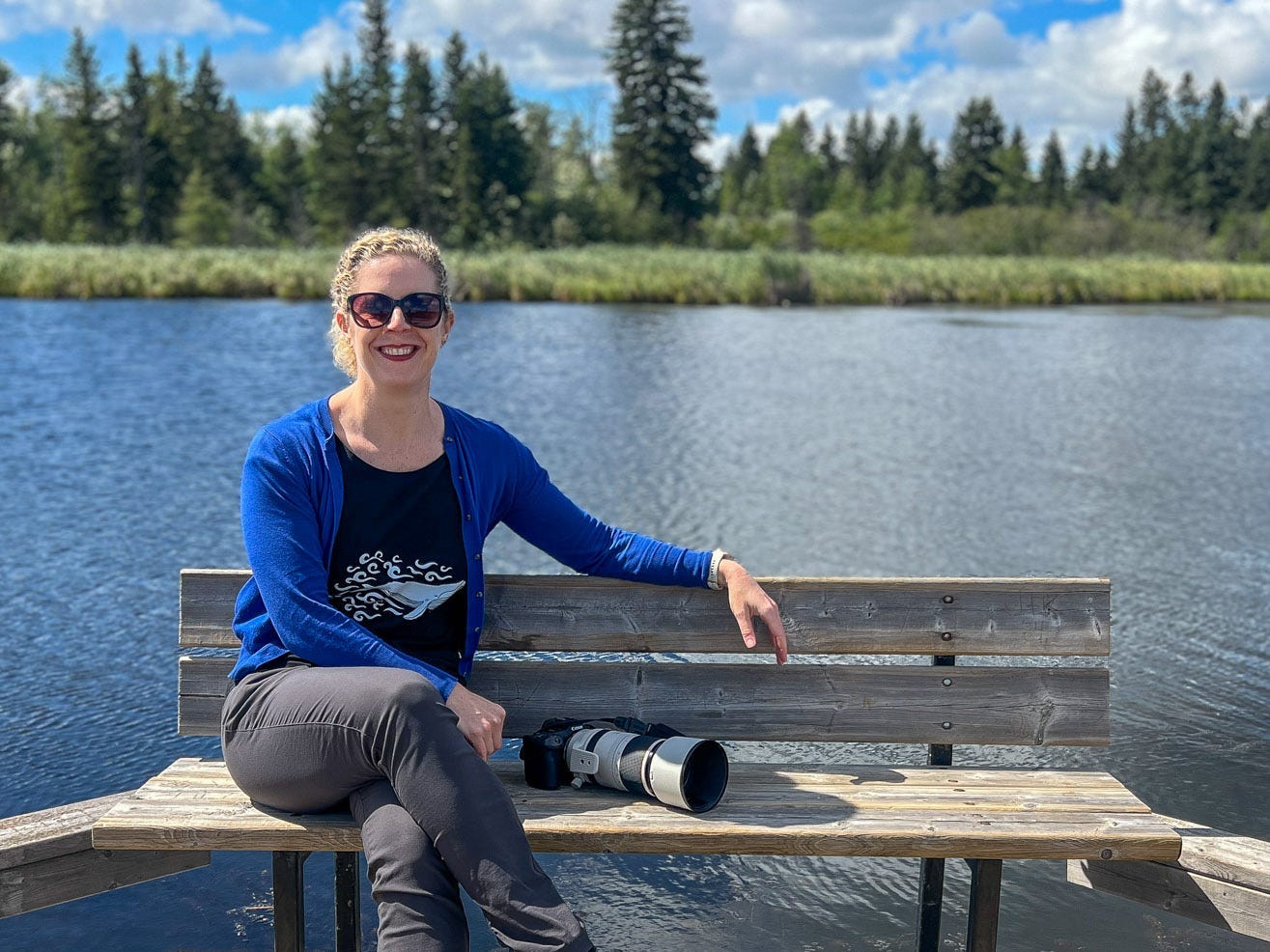 A woman wearing sunglasses is sitting on a bench next to a camera, with a lake in the background.