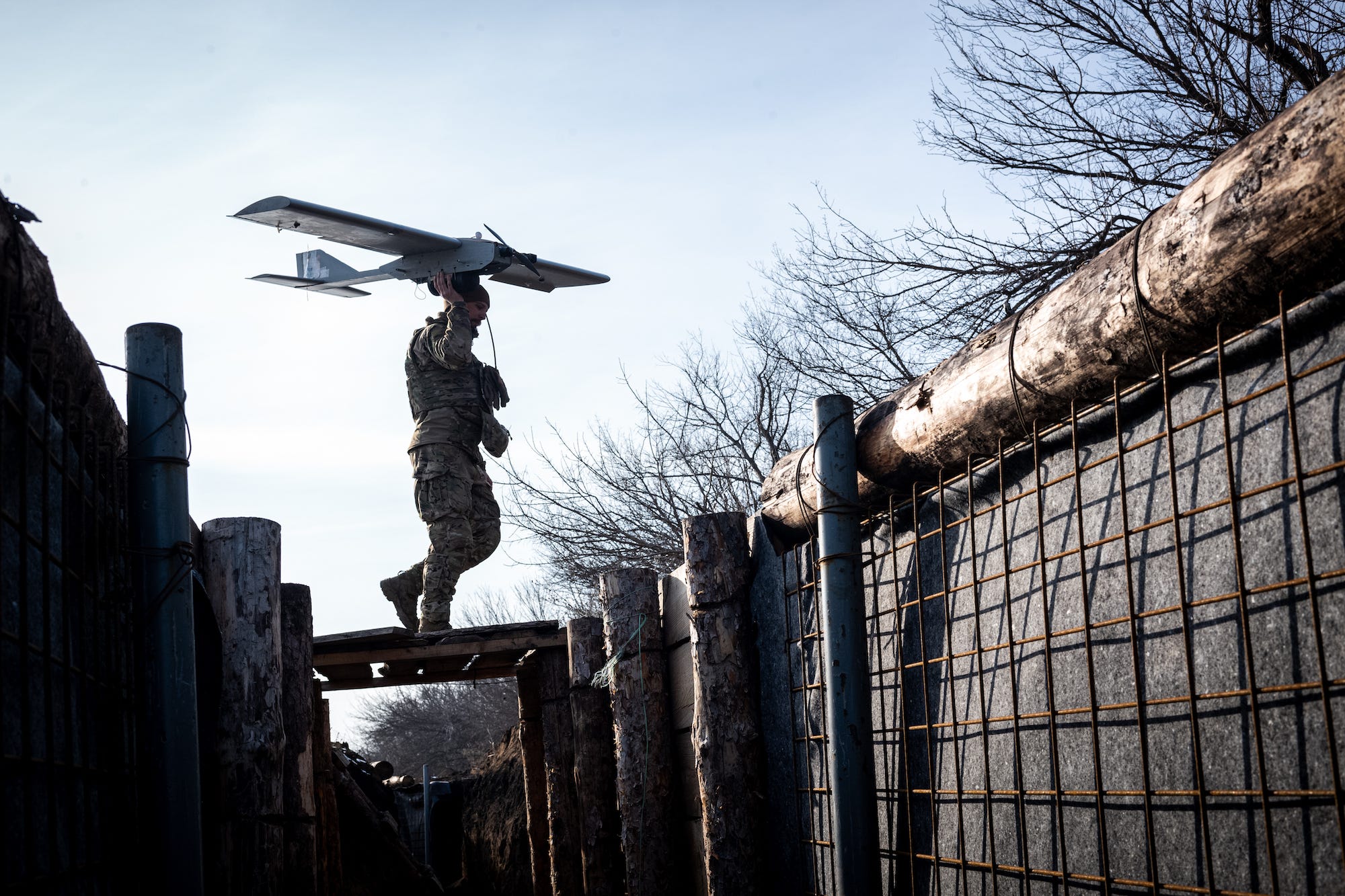 A man in combat dress holds a large drone in the air as he walks over planks of wood over a trench