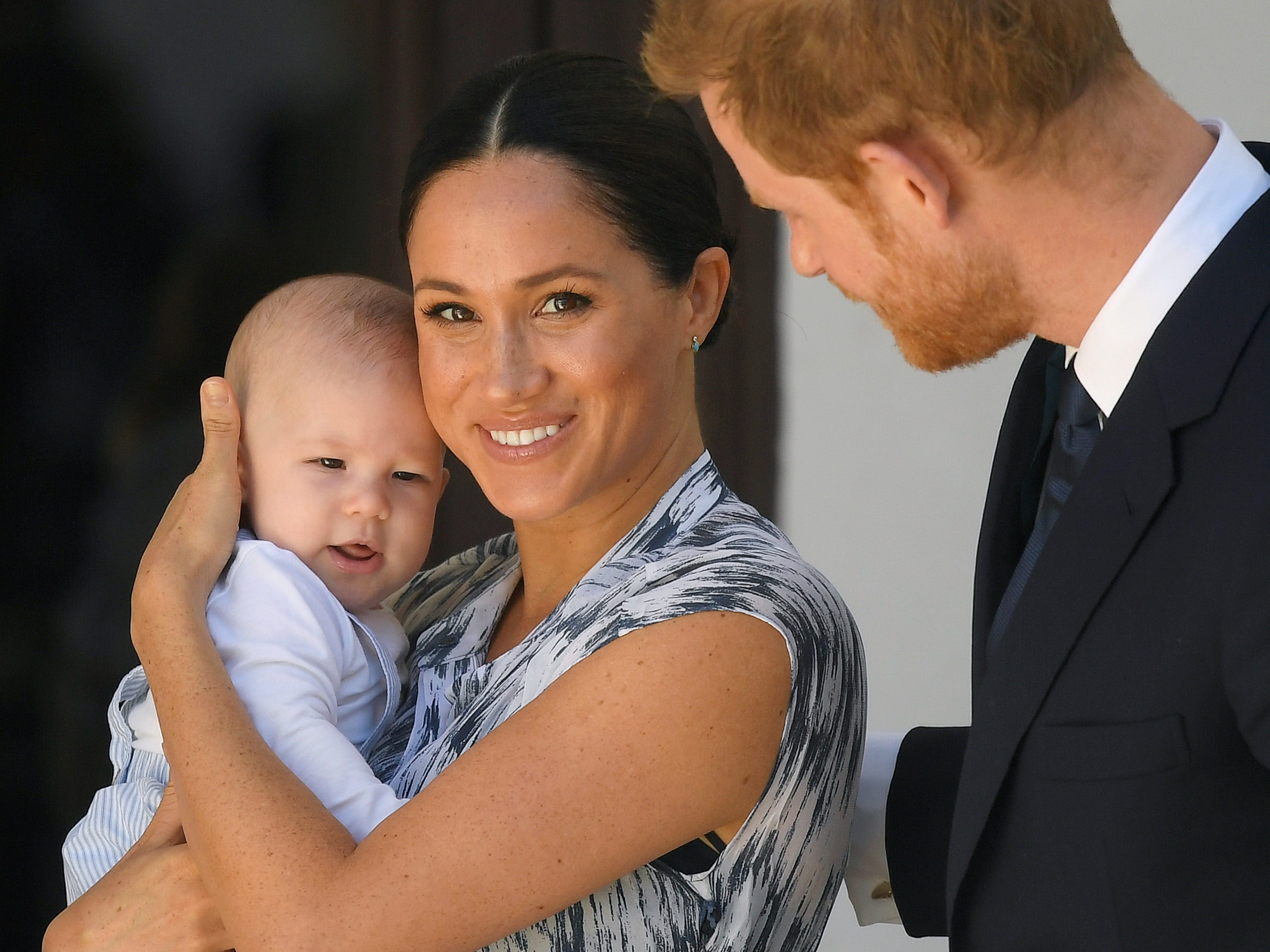 Meghan Markle looks at the camera while holding her first child, Archie, as Prince Harry touches her back.