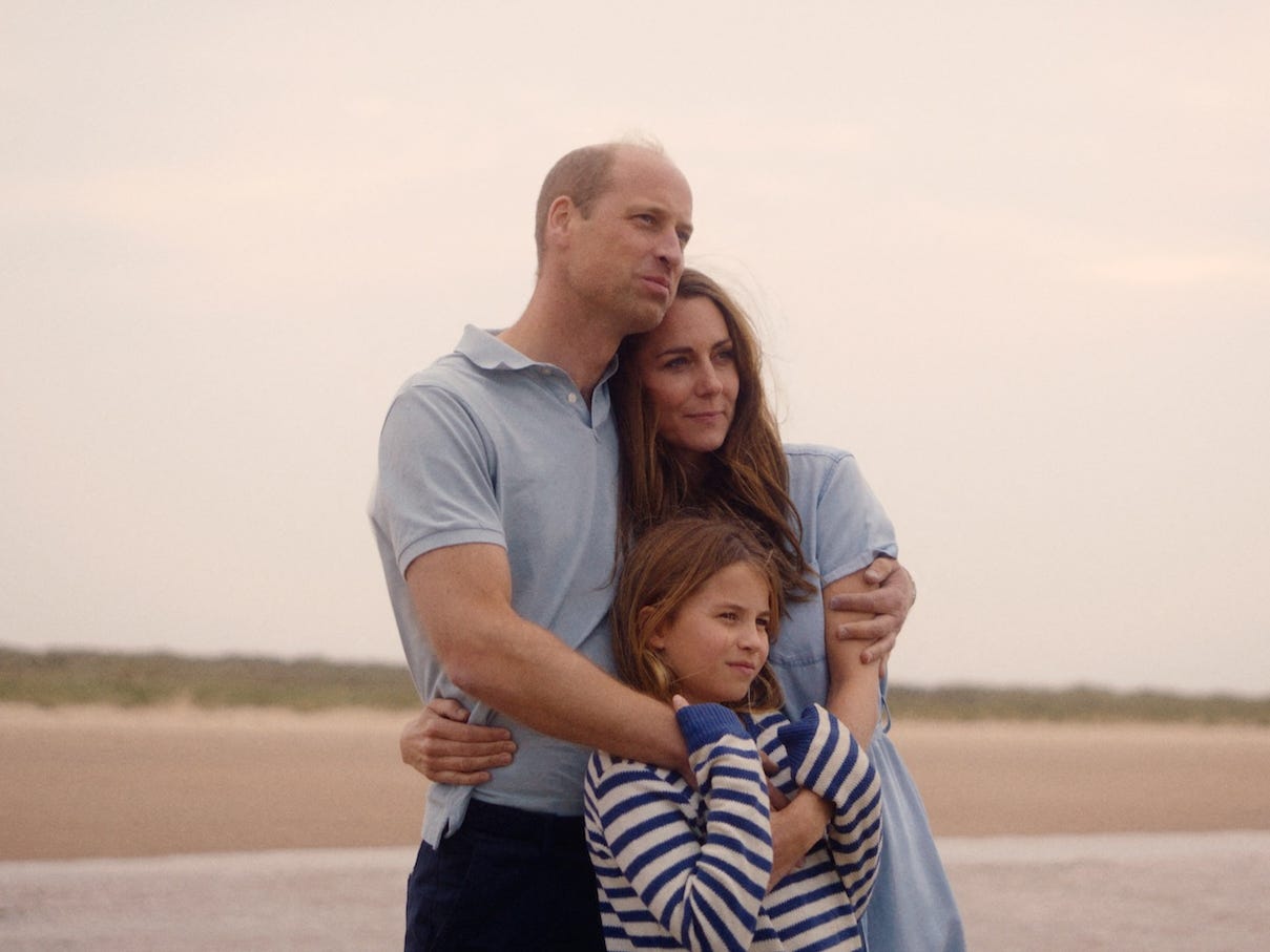 Prince William, Kate Middleton, and Princess Charlotte hugging on a beach in September 2024. William is dressed in a blue polo, Kate in a blue dress, and Charlotte in a blue and white striped long-sleeve shirt.