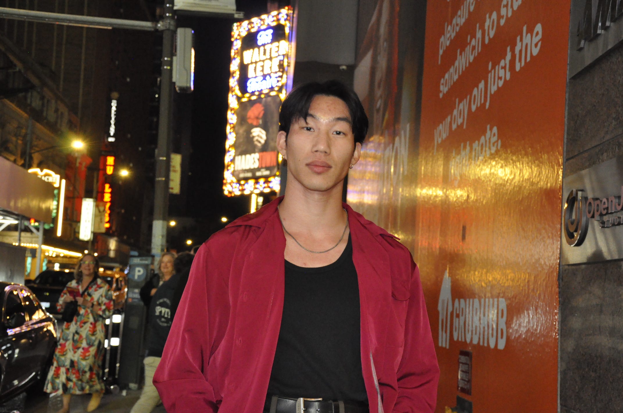 Aaron Yang, 22, stands in front of a neon billboard wearing a red jacket and black tank top.