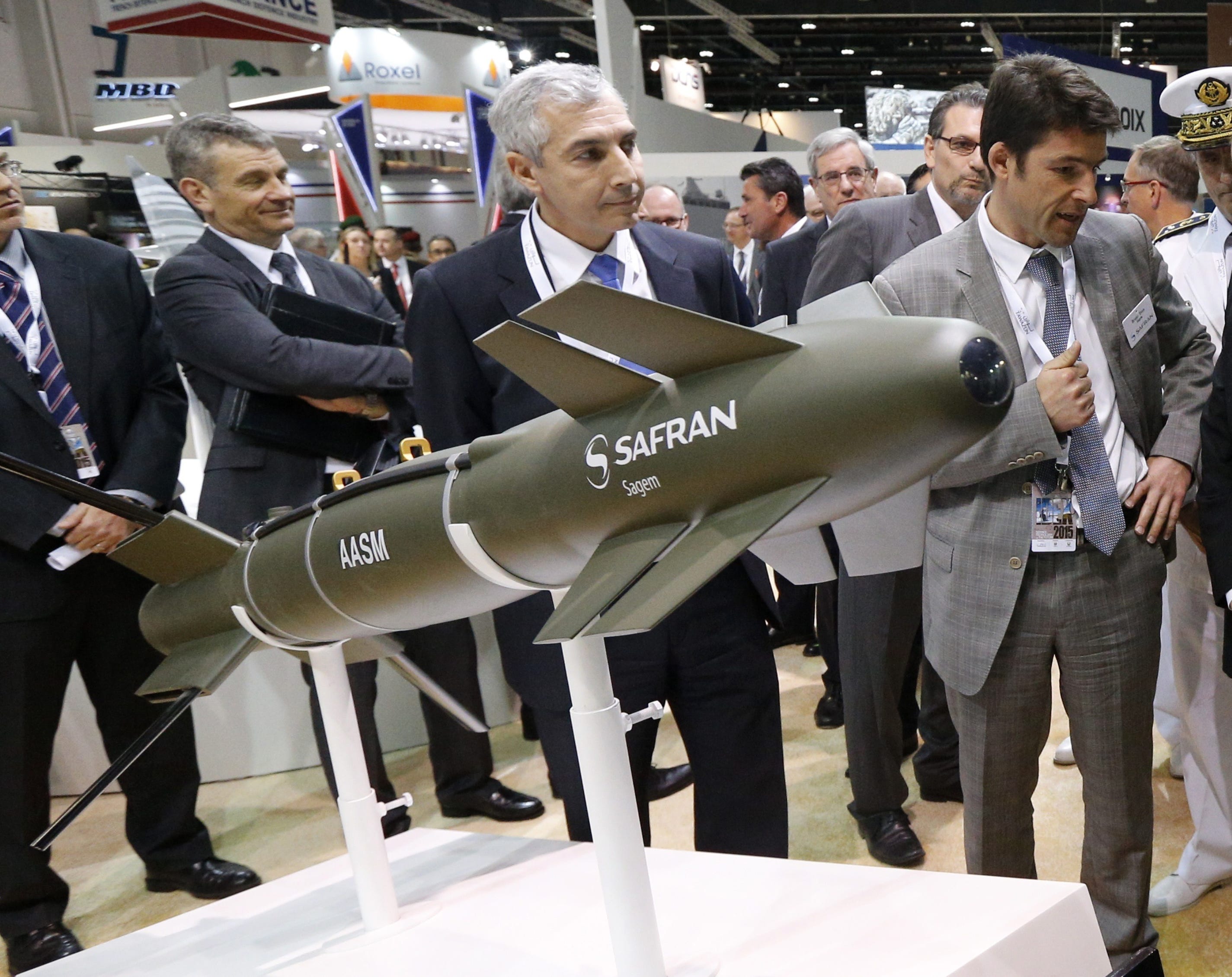 Men in suits stand around a green, cylinder-shaped piece of weaponry on a white stand